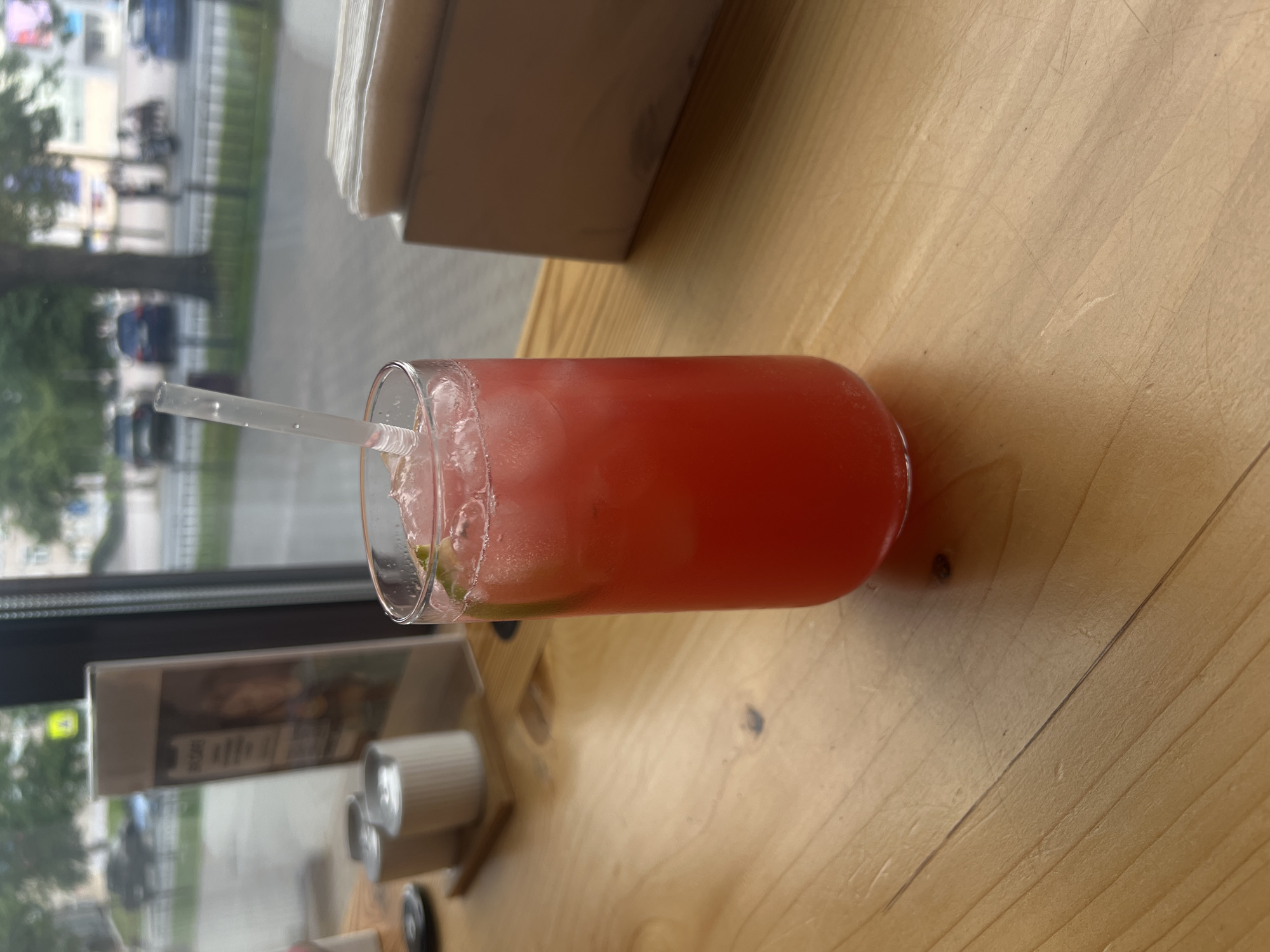 A tall glass of pink, icy liquid with a clear plastic straw is positioned on a light-colored wooden table. The drink appears to be a cold beverage, possibly a cocktail or juice, with ice cubes and a slice of lime visible near the rim. In the background, through a large window, a street scene is visible, suggesting the location is a cafe or restaurant. Cars and trees can be seen on the street, and a yellow traffic sign is visible in the distance. To the left of the drink, there are two small white cylindrical containers and a menu stand. To the right, a light-colored rectangular object, possibly a napkin dispenser, is present. The overall setting suggests a casual, urban environment in Chaikovsky, Russia.