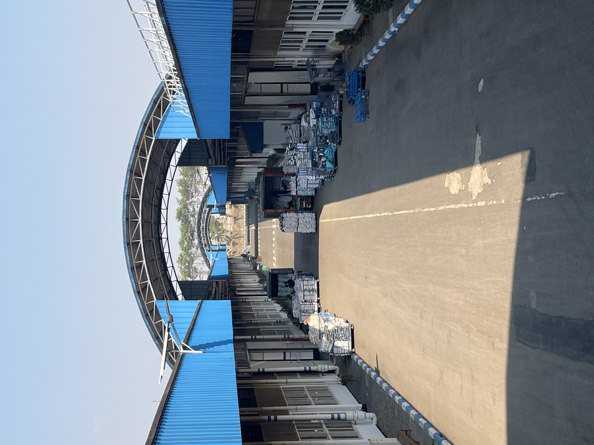 The image depicts an industrial facility in the city of Pelang, Indonesia, characterized by a long, covered walkway with blue corrugated metal siding. The walkway extends into the distance, forming a tunnel-like structure over an asphalt road. On either side of the road, there are rows of stacked goods, predominantly packaged in white sacks and plastic wrap, arranged on pallets. Some of these pallets are on wheels, suggesting they are ready for transport.
Several vehicles are visible. In the mid-ground, a green truck is positioned with its back open, and individuals appear to be loading or unloading goods. Closer to the foreground, on the left, a person is visible near a stack of pallets. Further down the road, another truck is parked. The ground is an asphalt surface, with a white line marking the center of the road. The sky above is clear and bright blue, indicating a sunny day. The overall environment suggests an active logistics or manufacturing hub.