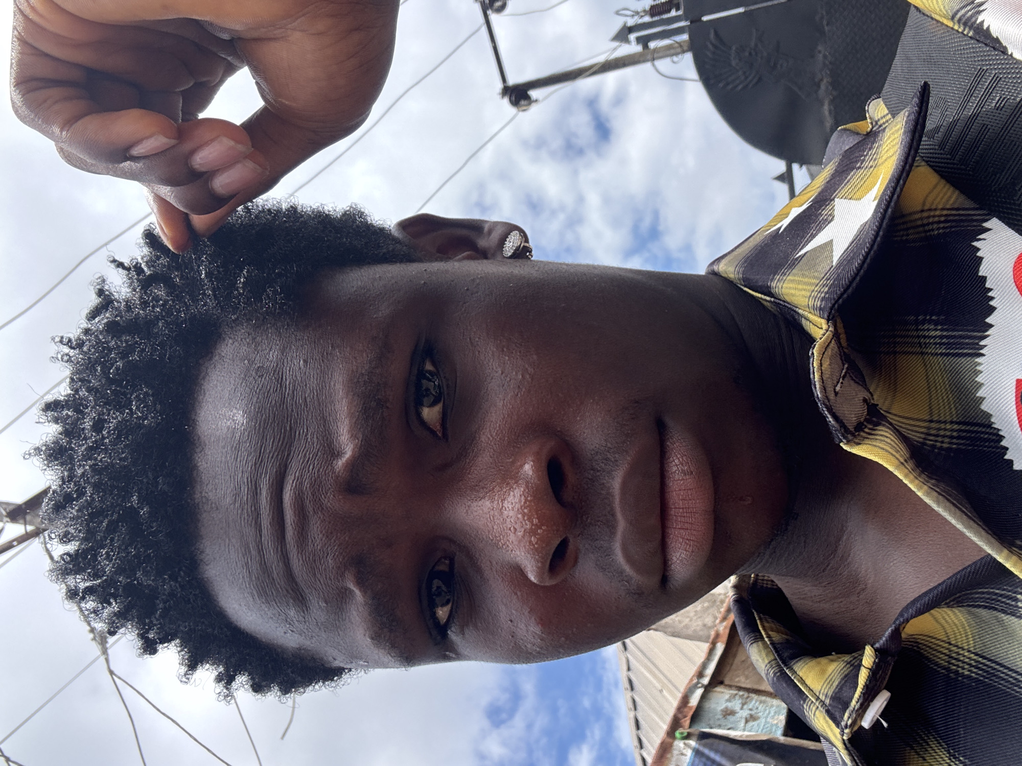 A young Black male with dark skin and short, curly hair is taking a selfie outdoors. He is wearing a black and yellow plaid shirt with white star patterns on the collar. He has a diamond stud earring in his left ear and is touching the top of his hair with his right hand. His expression is serious, with a furrowed brow. The background shows a cloudy sky with power lines and some corrugated metal structures, indicative of an urban or semi-urban environment. Based on the provided information, this scene is located in Ihiagwa, Nigeria.