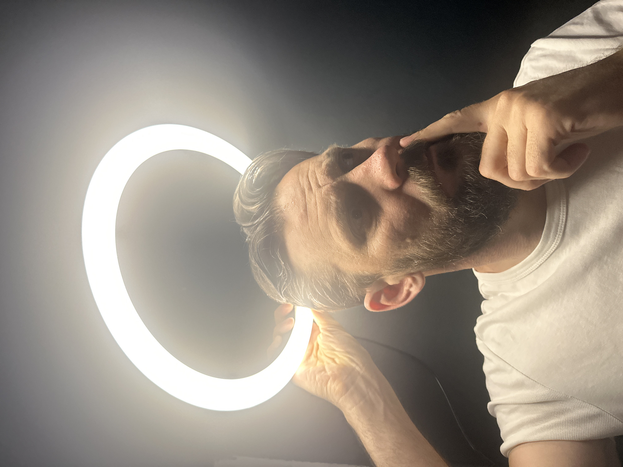 A close-up, diagonally composed shot features an adult man with a beard and greyish hair against a dark, undefined background. He wears a white, ribbed t-shirt. The primary light source is a brightly illuminated circular ring light, which he holds with his left hand, positioned near his head and chin. The intense light casts strong highlights on his hair, forehead, and beard, and illuminates his face directly. His right hand's index finger is pressed against his nose, while his eyebrows are furrowed, and his eyes are wide, conveying an intense, perhaps perplexed or thoughtful expression. The overall lighting suggests an indoor setting, likely in a dark room or at night, with the ring light being the dominant source of illumination. The image lacks visible text. This scene is contextualized as taking place in Fethiye, Turkey.