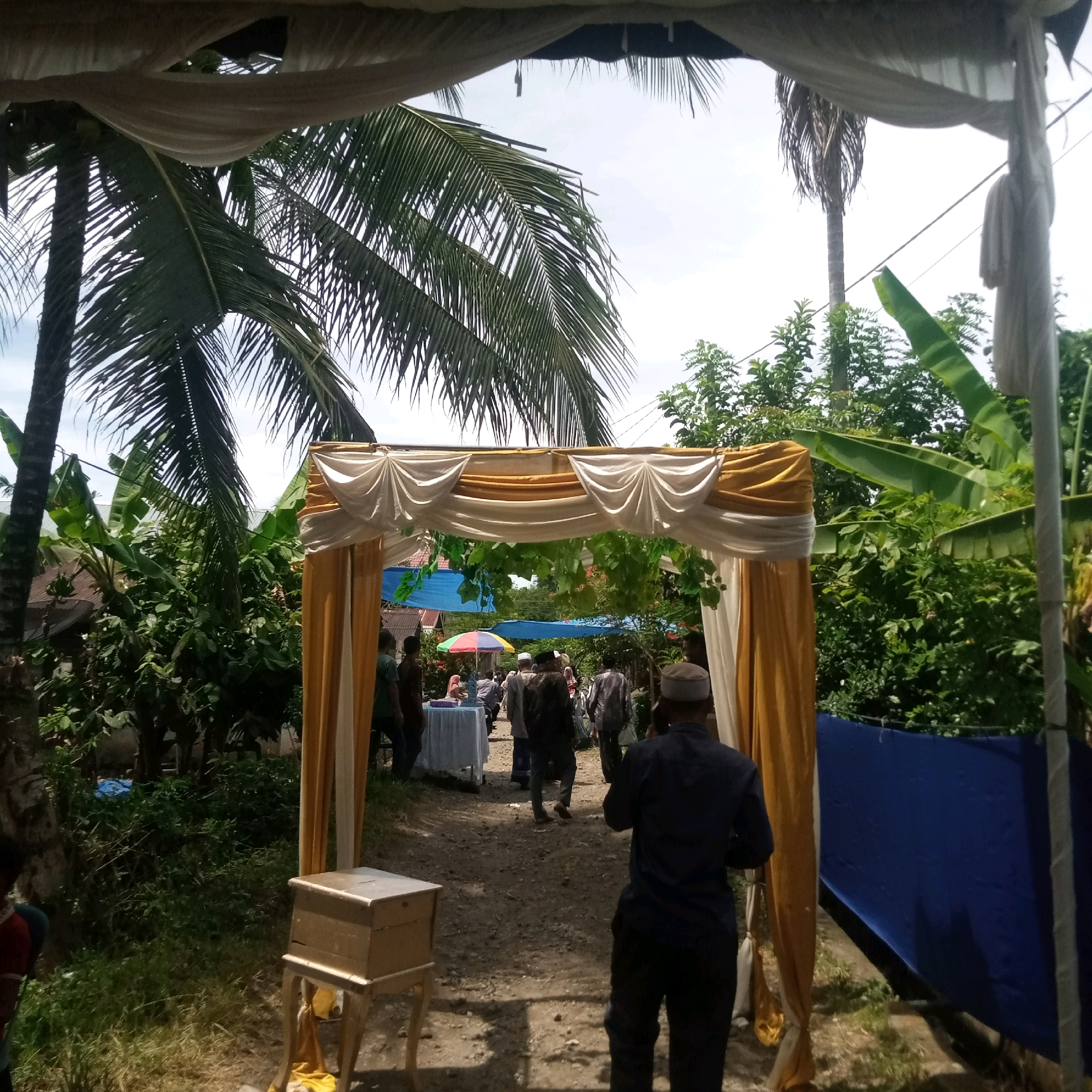 The image shows an outdoor wedding or event setup in Sareeaceh, Indonesia. A decorative archway draped with white and gold fabric spans across a dirt pathway. Several people are visible walking along the pathway and gathered near tables in the background. Palm trees and lush green foliage surround the area, suggesting a tropical or subtropical climate. There is a small, ornate wooden box on a stand to the left of the archway. A blue tarp is visible on the right side of the image. The sky is overcast.