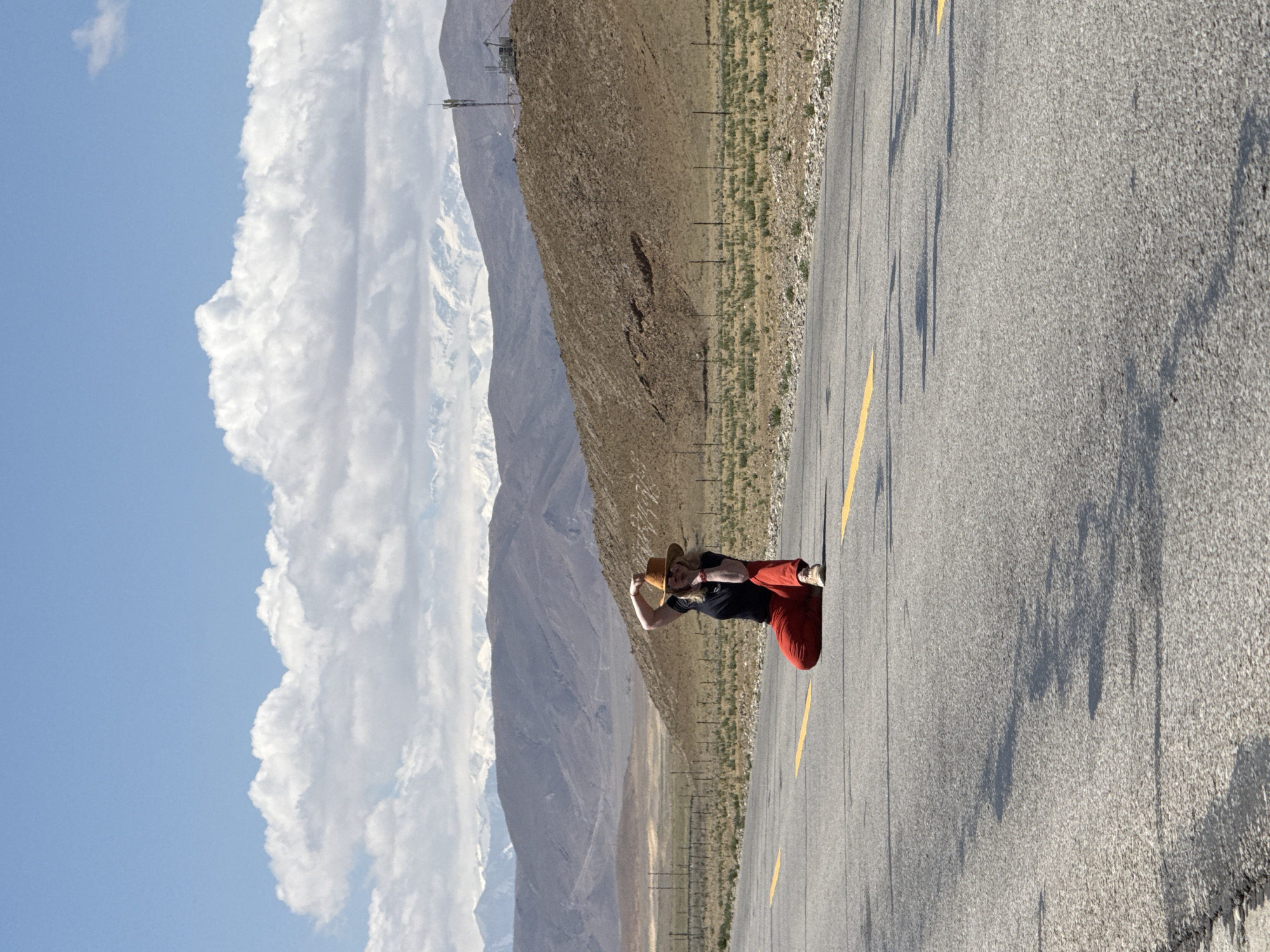The image depicts a vast, sunlit mountainous landscape, likely in Mainpu township, China. In the foreground, a two-lane asphalt road stretches into the distance with a single yellow dividing line. Long, distinct shadows are cast across the road from the left, suggesting it is daytime, possibly late morning or early afternoon.
A person stands in the mid-ground on the left side of the road, dressed in a black top, red pants, and a wide-brimmed straw hat. They are holding an object, possibly a camera or phone, up to their face, seemingly engaged in taking a picture or observing the scenery.
To the right of the road, a dry, undulating hillside covered in sparse, yellowish-brown vegetation and rocky terrain rises. A series of small posts, possibly utility or fence markers, are visible along this embankment. In the far background, towering mountain ranges dominate the horizon, with some peaks distinctly capped with snow or ice. The sky above is a vibrant blue, filled with large, fluffy white cumulus clouds. The overall scene conveys a sense of serene natural beauty and expansiveness in a high-altitude environment. There is no visible text in the image.
