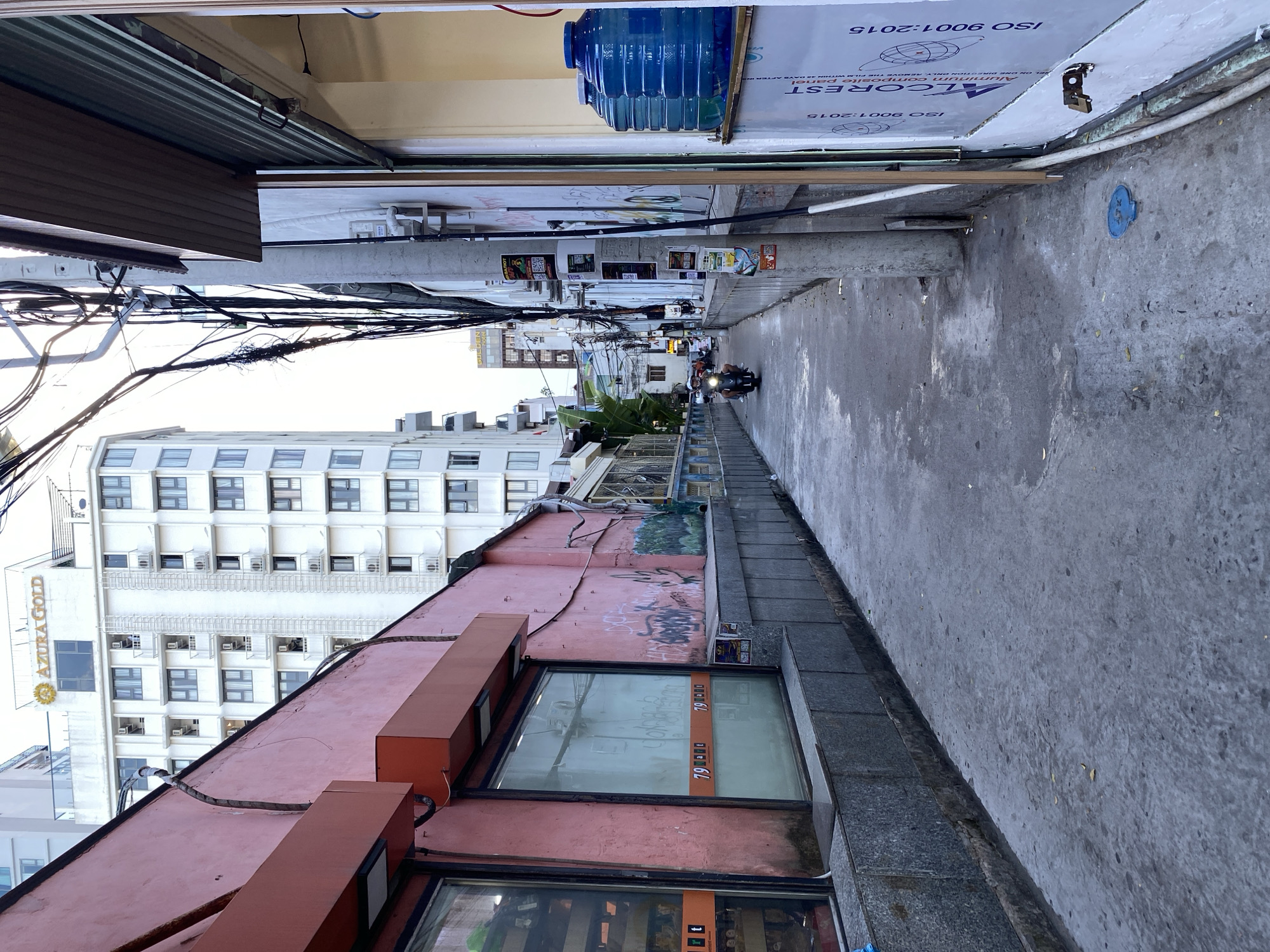 The image depicts a narrow alleyway in Nha Trang Ward, Vietnam, with buildings on either side. On the left, a pink building features large storefront windows displaying goods, one with orange signage indicating "79 000." Graffiti is visible on the pink wall. A gray, textured wall runs along the base of the pink building. On the right, a white building has a metallic roll-up shutter partially closed. Large blue water jugs are stacked on a ledge near the shuttered storefront. The alleyway is paved with concrete and shows signs of wear. Overhead, a complex network of electrical wires and cables is strung between poles. In the distance, a person is riding a motorcycle down the alley. A tall, white building with many windows, labeled "AZURA GOLD" at the top, is prominent in the background on the left. The sky is overcast.