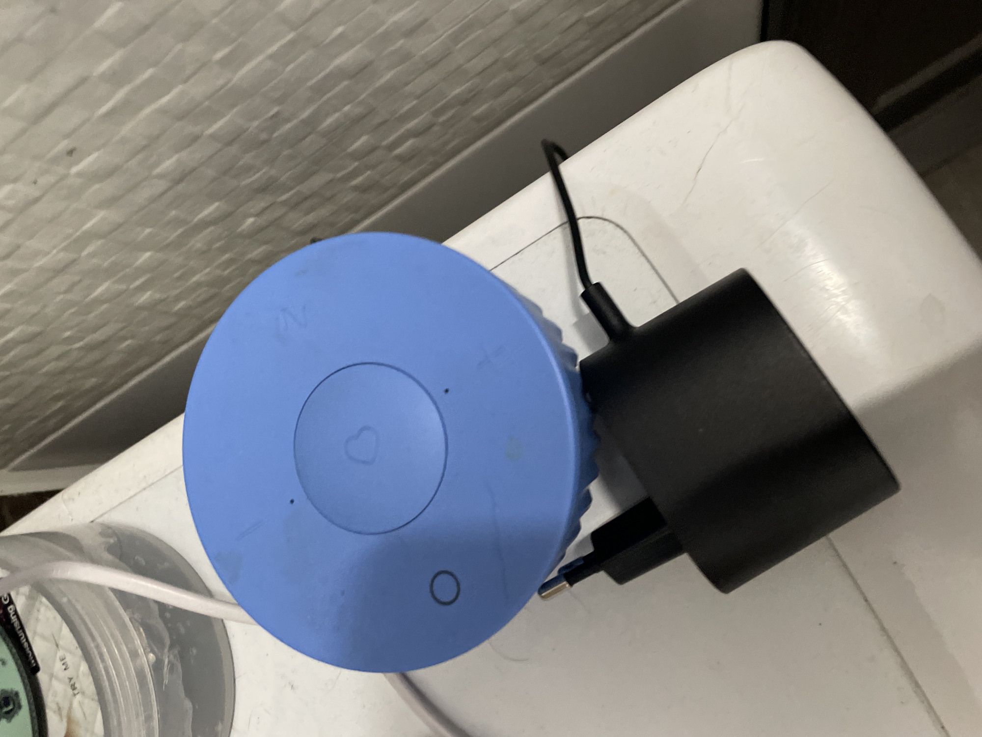 A blue electronic device with a heart symbol and a circle symbol on its top is connected to a black electrical adapter. The device and adapter are placed on a white surface. In the background, a textured grey wall is visible. The scene is photographed from a high angle. The location is the city of Sinyavino, Russia.