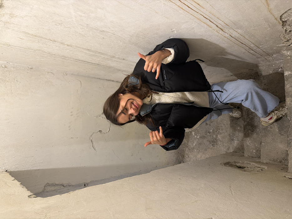 The image displays a young adult female, appearing to be in her 20s, standing on a concrete staircase within an indoor environment. The specific location within Tel-Aviv, Israel, cannot be identified from the provided visual details.
The female subject is positioned slightly below the camera's vantage point, looking upwards and smiling broadly with squinted eyes. Her hair is dark brown, worn down and wavy. She is dressed in a black, cropped puffer jacket with a visible white or cream lining around the collar and cuffs; a "PRADA MILANO" triangular logo patch is affixed to the left chest area. Underneath, she wears a cream-colored cable-knit sweater. Her lower attire consists of light blue, wide-leg corduroy pants with a visible drawstring, and light-colored, worn athletic shoes. She is performing a "shaka" hand gesture with both hands, thumbs and pinky fingers extended. A thin ring is visible on her right index finger, and a subtle red string or bracelet on her left wrist.
The environment is a narrow stairwell composed of concrete steps and unpainted, rough-textured walls. The walls show significant signs of wear, including multiple cracks, chipped plaster, and vertical brown streaking marks, particularly on the right wall. A prominent irregular indentation or hole is visible in the left wall towards the bottom of the frame. The concrete steps are worn and appear somewhat soiled. Lighting is diffuse, consistent with an interior setting, and highlights the rough textures and imperfections of the surfaces.