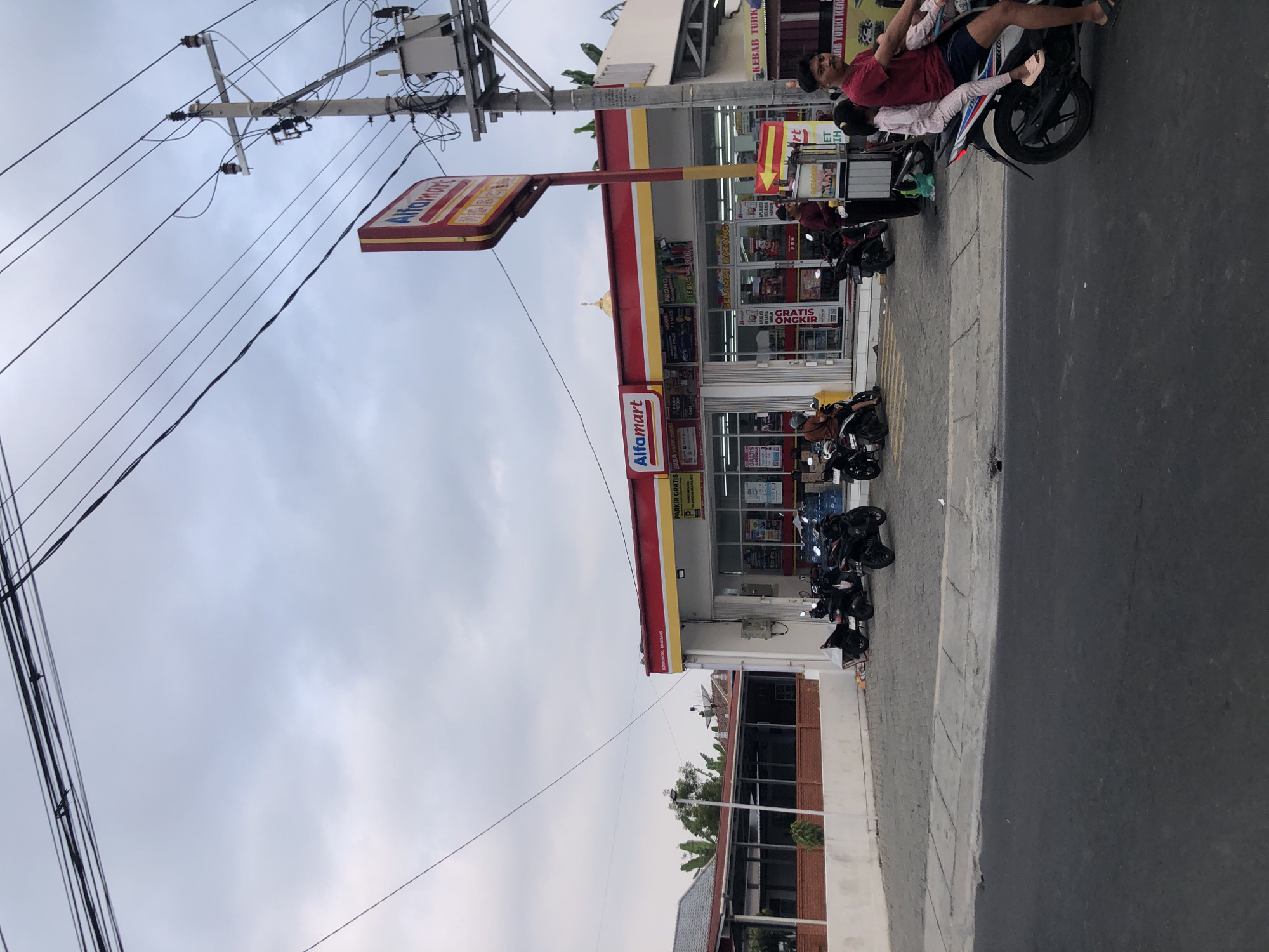 A street-level, slightly angled medium shot captures an Alfamart convenience store in Bondowoso, Indonesia, under an overcast sky. The store, with its prominent red and yellow signage, is the central focus. Several motorcycles are parked in front of the store, and one is on the road in the foreground, carrying a man and two young children. A man stands at a food cart adjacent to the store, and other individuals are visible inside the store. The environment includes a sidewalk, a paved area, a road, a building to the left with red brick accents, and utility poles with numerous wires overhead. The overall scene depicts a typical commercial street with everyday activity.