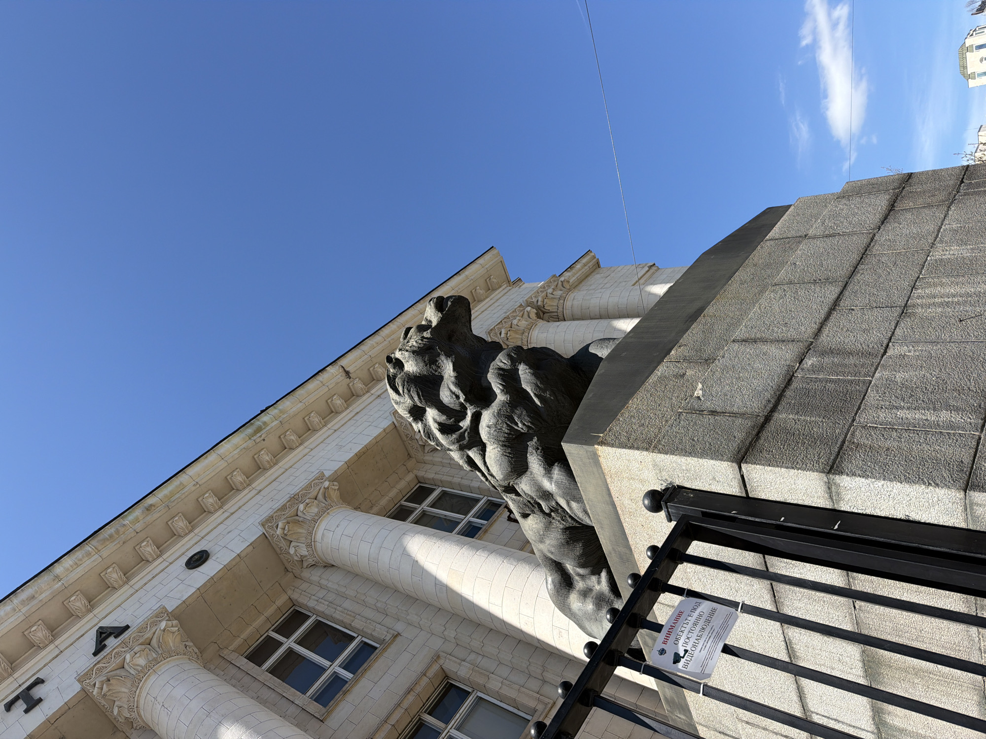 The image captures a low-angle view of a building and its sculptural elements in Sofia, Bulgaria. A weathered lion sculpture, presumably made of stone, dominates the frame, situated atop a large, stepped stone structure. Adjacent to the sculpture, a building with classical architectural features is visible, including columns, windows, and decorative elements. A black metal fence is in the foreground, with a warning sign attached. The sky is bright blue with a few scattered clouds. The location is likely near Sofia University, identifiable by the building's architectural style and the lion sculpture.