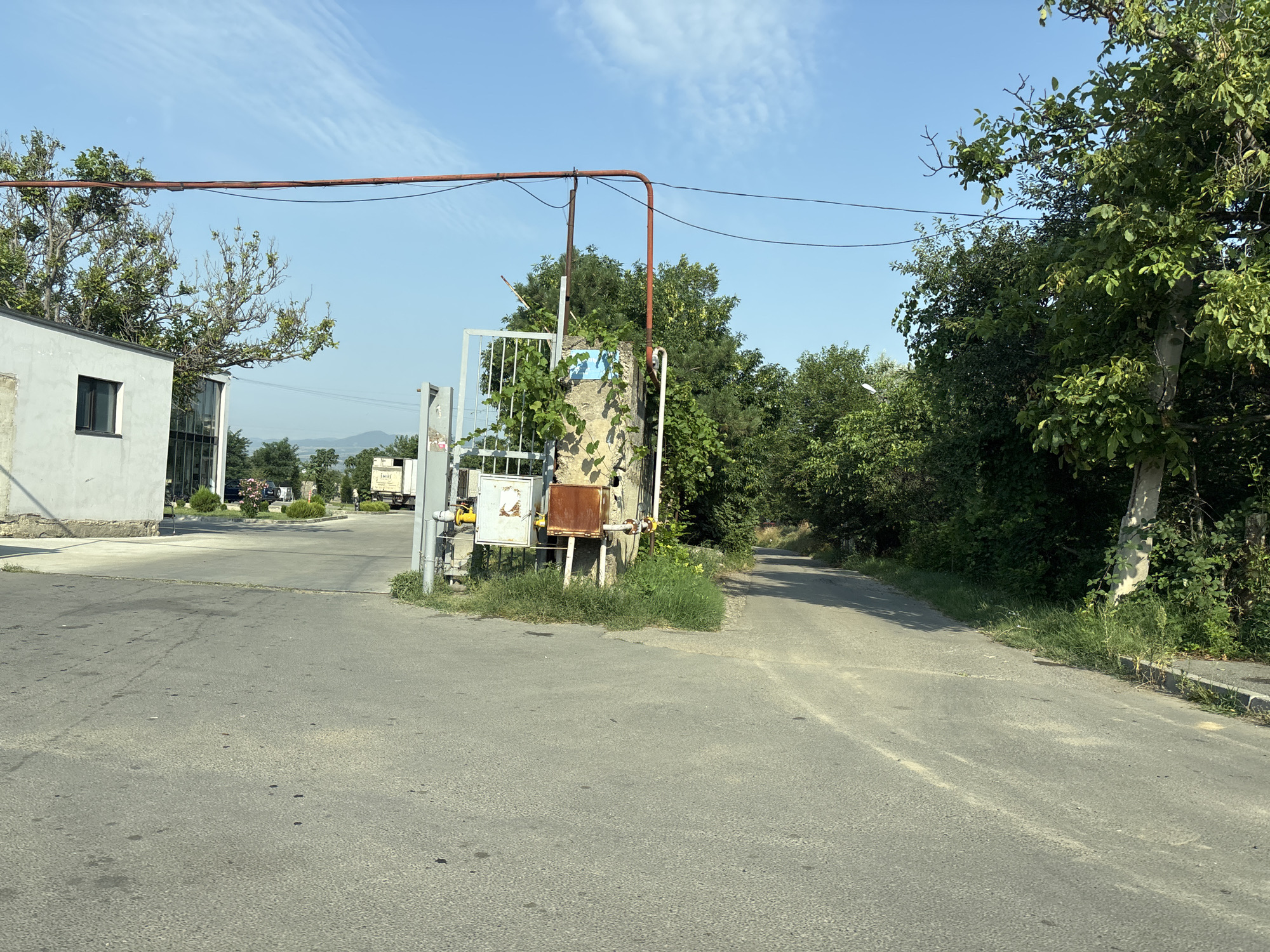 The image depicts an outdoor scene on a sunny day, likely in Tbilisi, Georgia. In the foreground, a paved road curves to the right. To the left, a light-colored building with a dark-trimmed window is visible. Adjacent to the building is a metal gate and a concrete structure covered in vines, with what appears to be utility boxes attached. A red pipe runs horizontally across the top of the frame, supported by poles. Behind the gate and concrete structure, a dense arrangement of green trees and foliage forms a backdrop. Further in the distance, the road continues into a wooded area, and the hazy outlines of hills or mountains are visible on the horizon. The sky is blue with scattered white clouds. There are no people or animals visible in the image. The scene suggests an entrance to a property or industrial area.