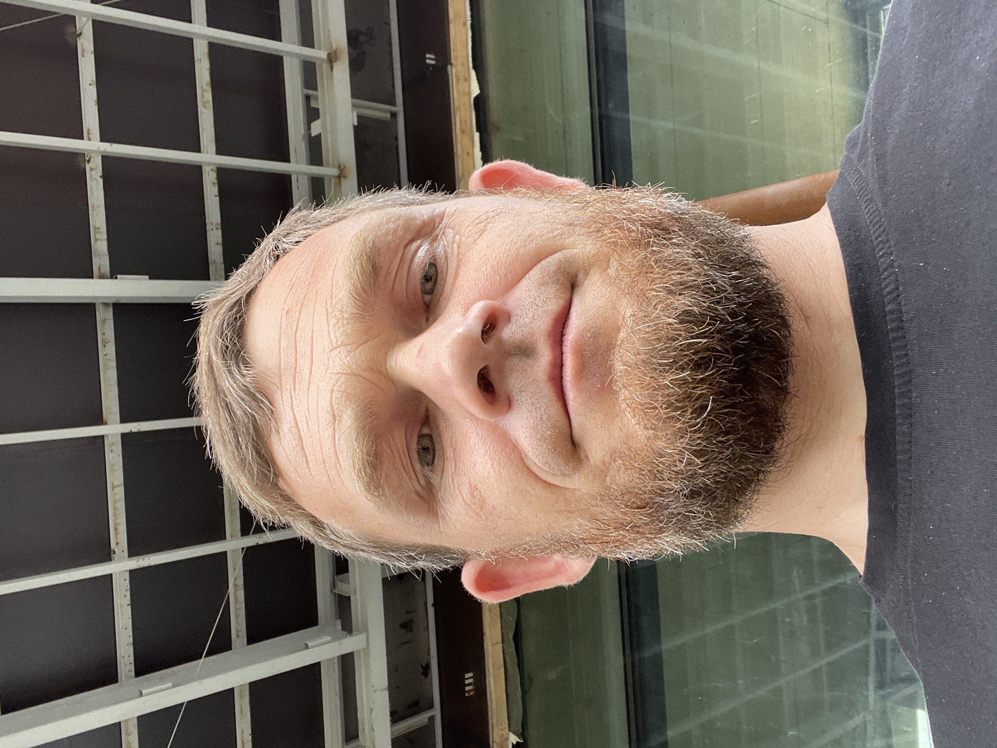 The image is a close-up, low-angle selfie of a man with light brown hair and a beard, wearing a black t-shirt. He has light skin with visible pores and wrinkles on his forehead. His eyes are looking directly at the camera. The background appears to be an outdoor construction site, with a metal frame structure and dark panels overhead, and a green reflective surface that could be a building facade or window. The specific location in Krasnodar, Russia, cannot be determined from the image alone.