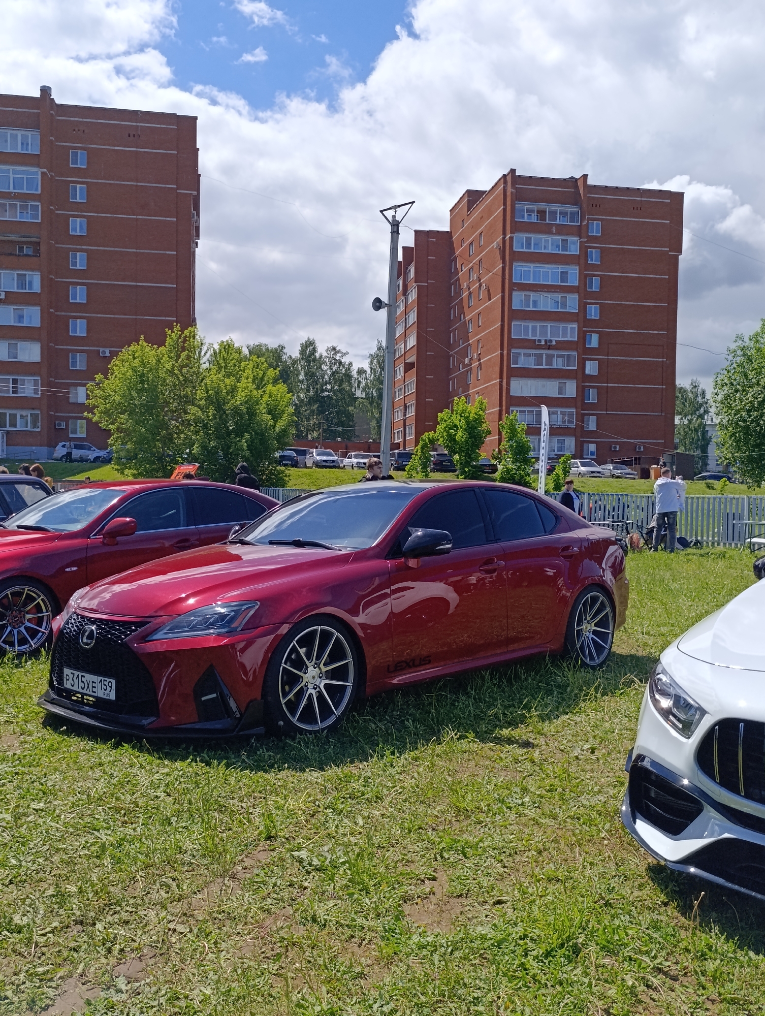 The image depicts a car exhibition in Chaikovsky, Russia, on a sunny day. The foreground features a maroon Lexus IS with a visible Russian license plate "P315XE 159" parked on a grass field. Another maroon Lexus is partially visible behind the first one, and the front of a white car is visible to the right. In the midground, a white picket fence separates the cars from a group of people, mostly standing around. Behind them, there are parked cars and some greenery. In the background, there are two large, brick apartment buildings with multiple windows and a street light between them. The sky is blue with scattered white clouds.