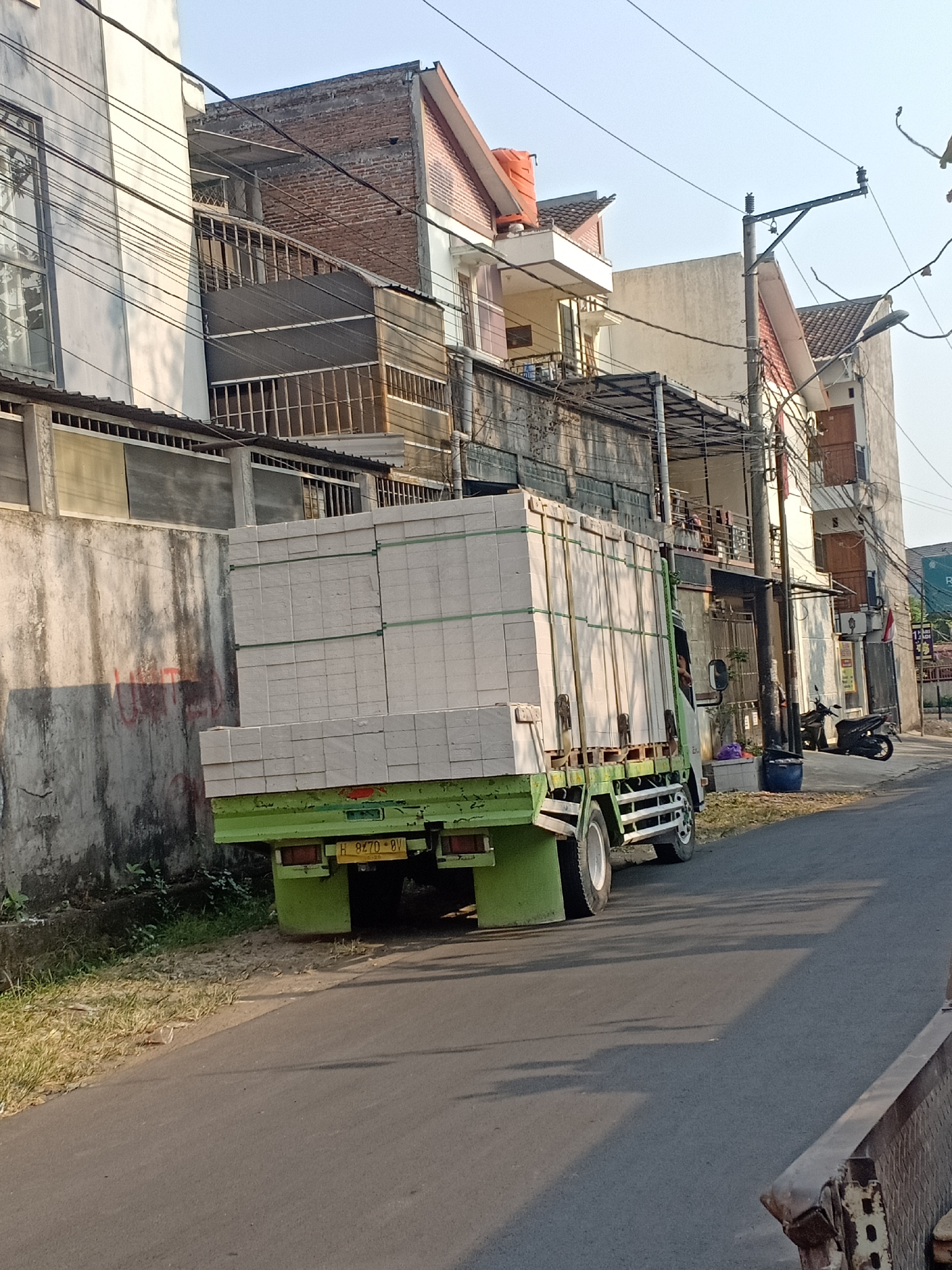 A light green truck is parked on the right side of a paved street in a residential area of Semarang, Indonesia. The truck is loaded with stacks of white, rectangular objects, likely building materials, secured with green straps. The truck's license plate, partially visible, reads "H 8270 BV". To the left of the truck is a weathered concrete wall with some red graffiti. Behind the wall, and on the right side of the street, are multi-story buildings with exposed brick, balconies, and satellite dishes. A black motorcycle is parked near the curb in front of one of the buildings. Overhead, a tangle of electrical wires crisscrosses the sky, supported by concrete poles. The sun is casting long shadows across the street, indicating it is likely late afternoon or early morning. No people are clearly visible in the image.
