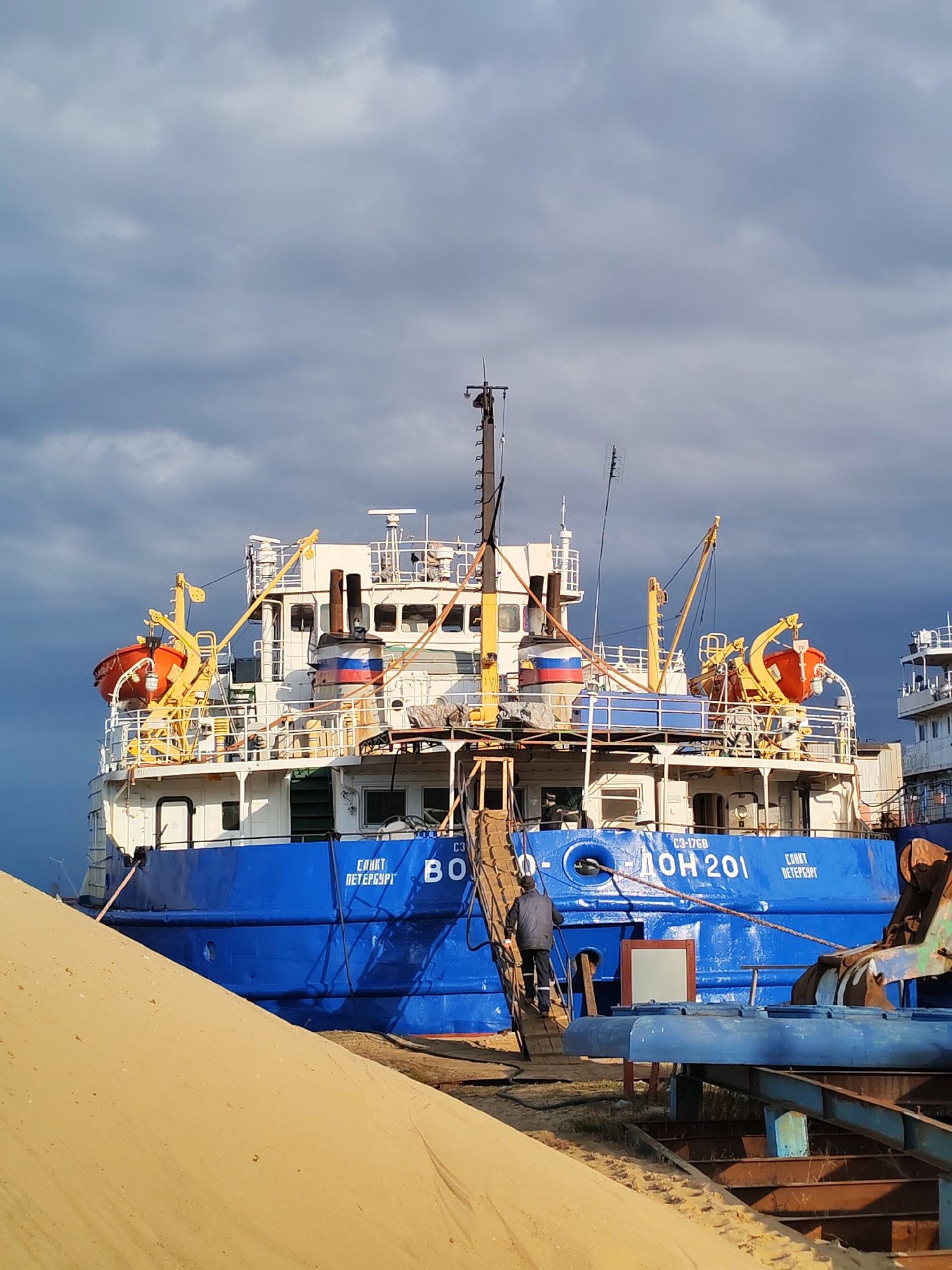 The image shows a blue and white ship, identified as "DON 201," docked in what appears to be Rybinsk, Russia, judging by the Cyrillic text on the ship "Санкт Петербург" (Saint Petersburg). A person in dark clothing is ascending a wooden gangway onto the ship. The ship has lifeboats, masts, and antennas. In the foreground, there is a large pile of sand. The sky is overcast with dark, looming clouds. Other industrial equipment and possibly another ship are visible partially in the background.
