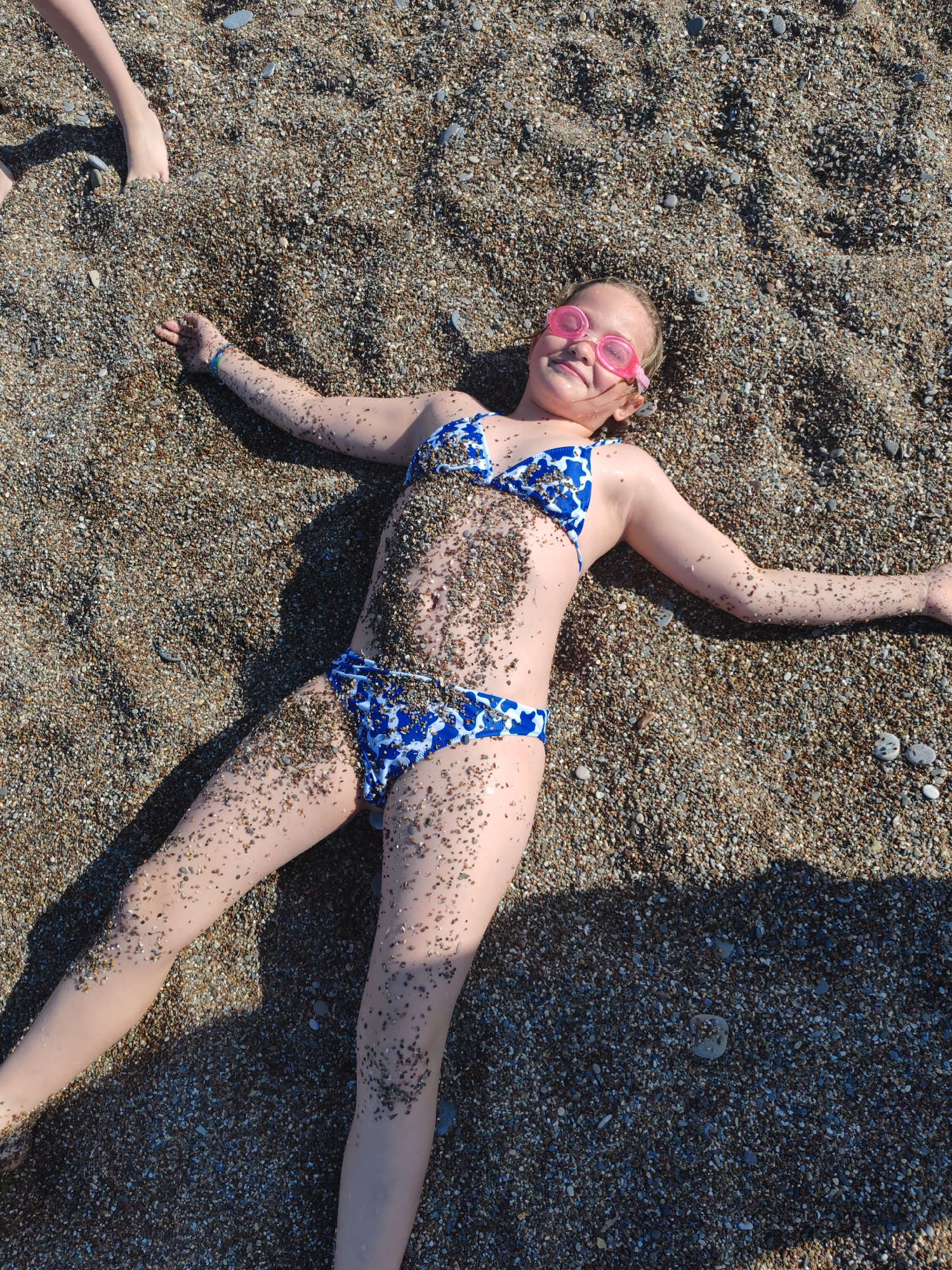 A young girl, estimated to be between 5-10 years old, is depicted lying on a beach covered in coarse sand and small pebbles. She is positioned centrally in the frame, lying on her back with her arms and legs spread out, forming a relaxed, star-like pose. She is wearing a blue and white patterned two-piece bikini and pink swimming goggles pushed up onto her forehead, covering her eyes. Her skin is fair, and her body, particularly her torso, arms, and legs, is speckled with small grains of sand and tiny pebbles. She has a happy expression, with a broad smile on her face.
In the top-left corner of the image, the lower legs and feet of another person are partially visible, suggesting they are also on the beach nearby. The scene is brightly lit by natural sunlight, casting distinct shadows, indicating a clear, sunny day. The texture of the beach is prominent, composed of various shades of brown, grey, and white pebbles, consistent with a Mediterranean beach such as those found in Alanya, Turkey. There is no visible text in the image. The overall impression is one of leisure and enjoyment on a sunny beach day.