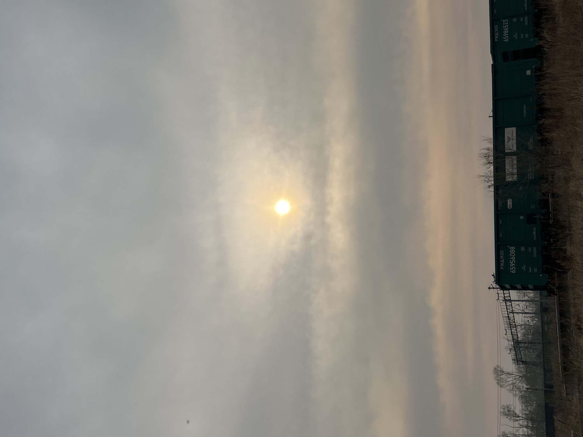 The image shows a hazy, overcast sky with the sun visible through the clouds. Adjacent to the sky are several green train cars. The location appears to be near a train yard or railway. The city is unknown. The scene lacks details about people or specific interactions.