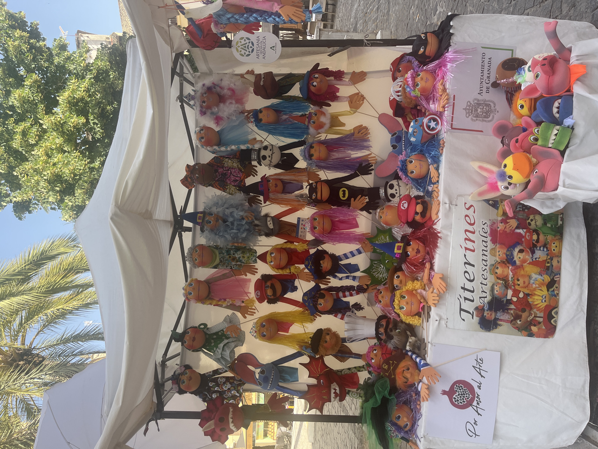 A stall displays a collection of handmade puppets and stuffed animals at an outdoor market in Granada, Spain. The stall is covered by a white canopy, and the ground is paved with cobblestones. The puppets vary in size and design, with some resembling characters from popular culture like Mario and Batman, while others are more whimsical creations. There are also several plush toys, including a pink pig, a yellow duck, and a blue shark. Signs on the stall advertise "Titerines Artesanales" (Artisan Puppets) and "Por Amor al Arte" (For the Love of Art), along with the official seal of the Ayuntamiento de Granada (City Council of Granada). Palm trees and lush green foliage are visible in the background, suggesting a warm, sunny day.