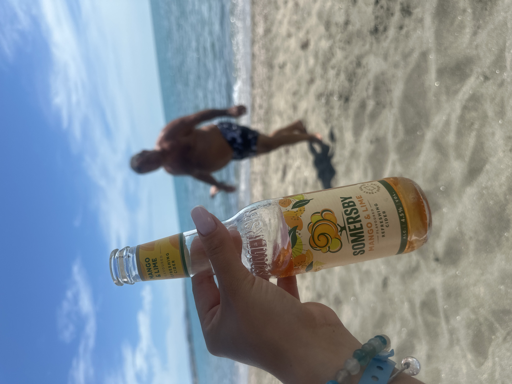 A hand is holding a bottle of Somersby Mango & Lime cider in the foreground. The bottle has a yellow label with an illustration of mangoes and limes, and text indicating the flavor and "Refreshing Cider". The hand has light-colored nail polish and is wearing a bracelet with blue and clear beads. In the background, a blurry figure of a man in swim trunks is walking on a sandy beach. The sea and a clear blue sky with some clouds are visible behind him. The scene is set on a beach in Aheloy, Bulgaria.