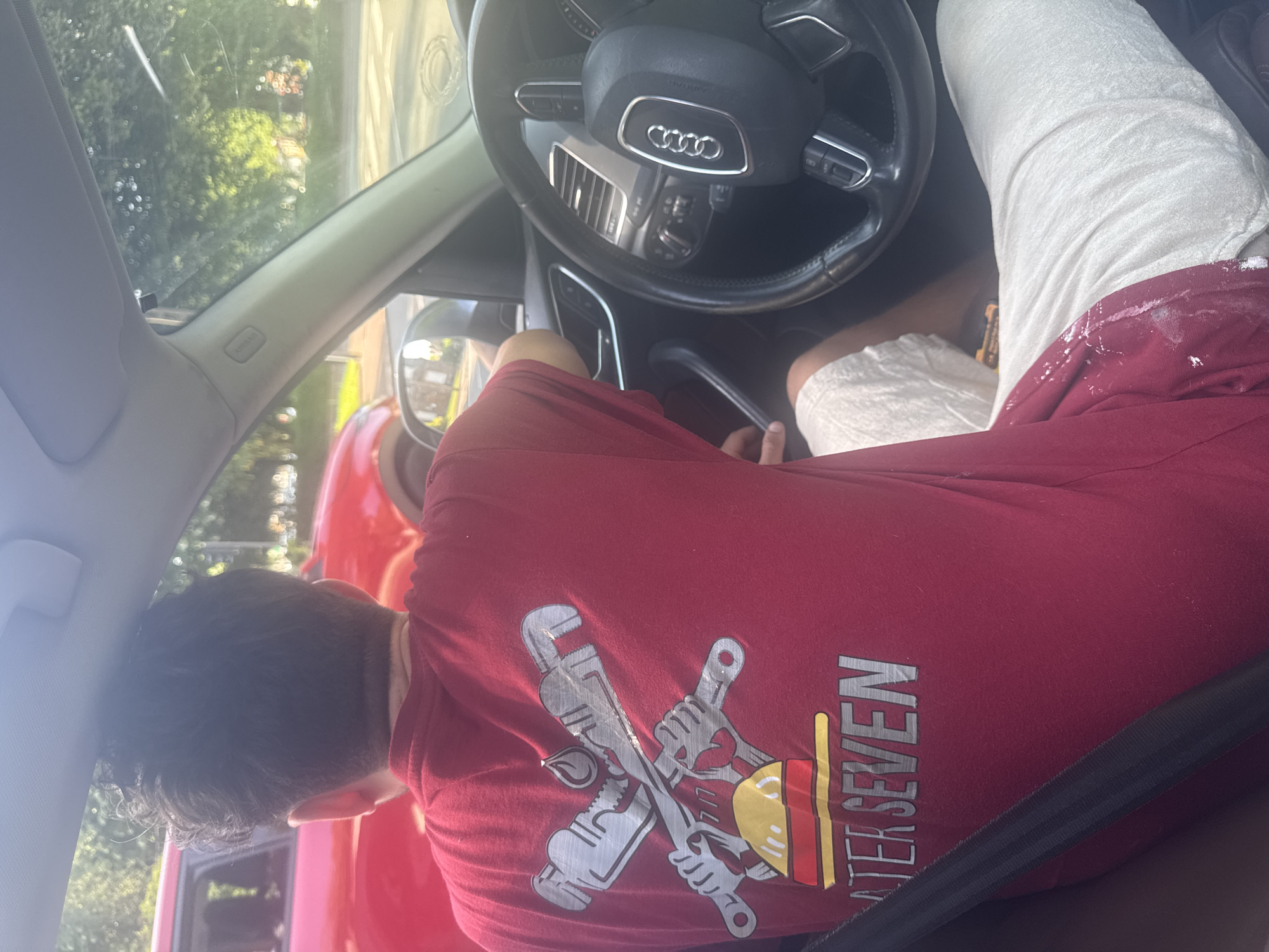 The image depicts a person in the driver's seat of an Audi. The steering wheel displays the Audi logo. The person is wearing a red t-shirt with a plumbing-related design on the back. Their legs are visible, with white shorts and a red stain, possibly paint, on one leg. The person is wearing a seatbelt. The location is Marcq-en-Barœul, France, based on the "Water Seven" t-shirt. The subject's head appears to be resting near the driver-side window.