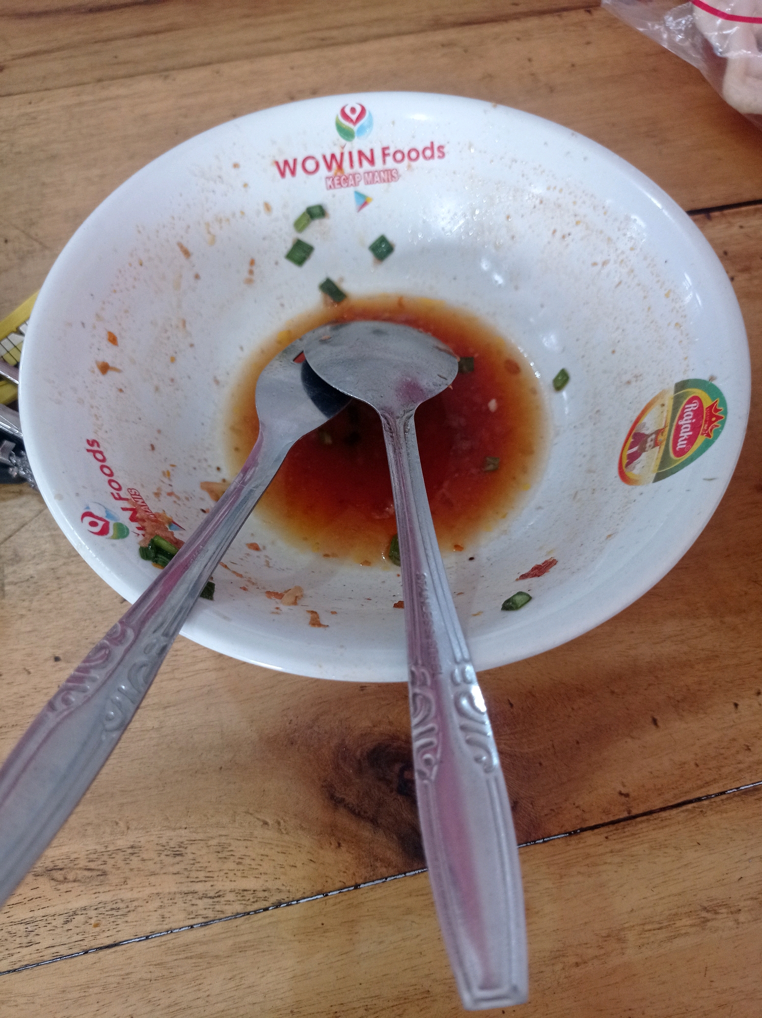 A high-angle shot shows a dirty white bowl with two spoons on a wooden table. The bowl contains some leftover red sauce, green herbs, and remnants of food. The text "WOWIN Foods" and "Kecap Manis" are visible on the inside of the bowl, indicating it is from a food establishment in Kedok, Indonesia. The spoons are made of stainless steel and have decorative patterns on their handles. The wooden table has a natural grain pattern and appears to be weathered. There is a plastic bag on the upper right corner of the image, partially obscuring the background.