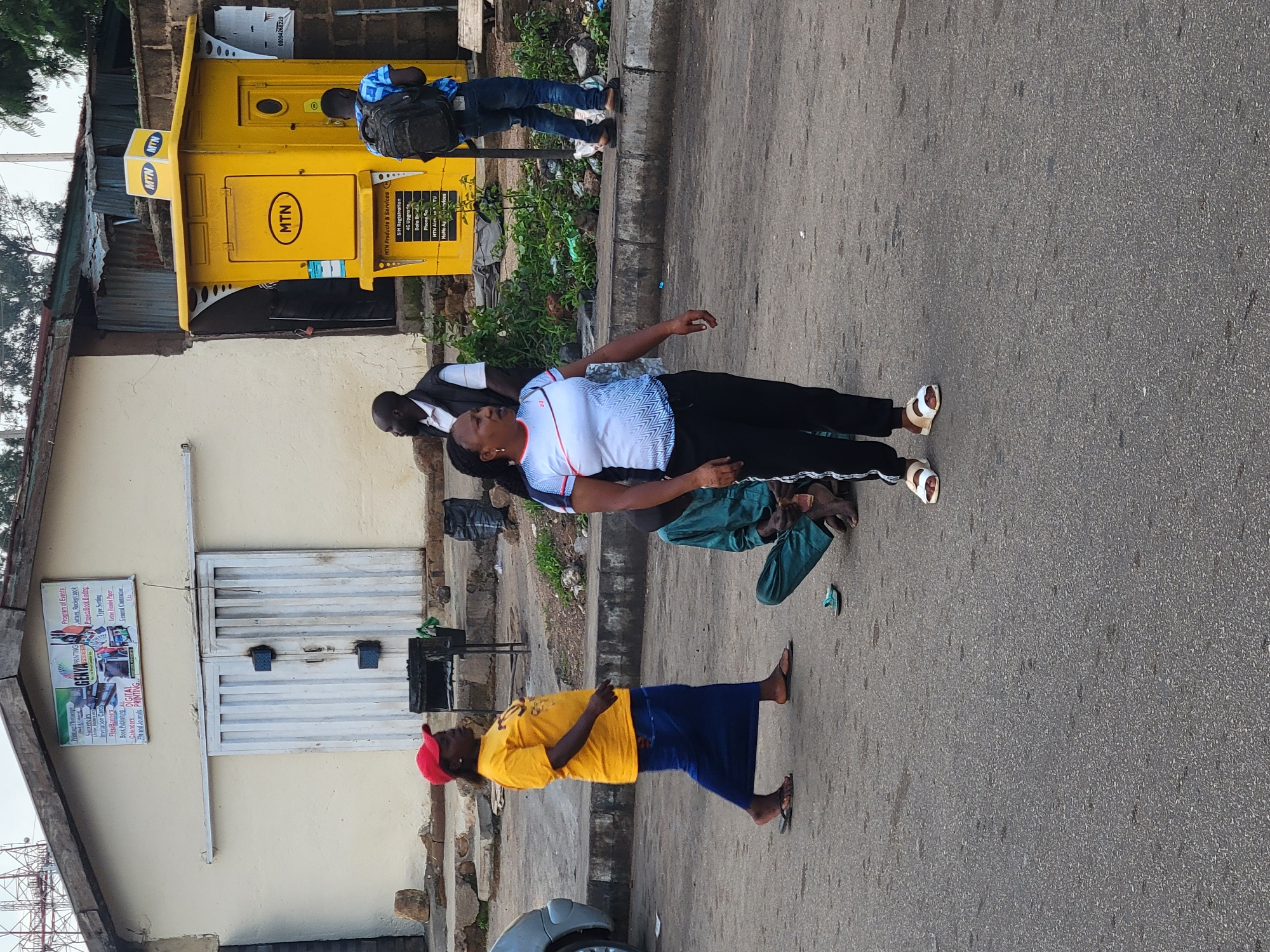 A group of people are gathered on a paved street in front of a yellow MTN kiosk and a building with a sign indicating printing services. In the foreground, a woman in a white and blue patterned t-shirt and black pants stands facing left, looking to her right. She is wearing white sandals. To her left, a woman in a yellow t-shirt and blue skirt walks past, wearing flip-flops and a red cap. A man in a green garment is crouched on the ground to the left of the woman in the white t-shirt, holding something in his hands. In the background, a man with a backpack walks away from the MTN kiosk, and another man stands near the entrance of the printing shop. The MTN kiosk is a bright yellow, multi-sided structure with the MTN logo prominently displayed. The building behind the kiosk is light-colored with a corrugated metal roof. The street appears to be in an urban or semi-urban setting, with some debris visible on the side of the road. The overall scene suggests a public space with commercial activity.