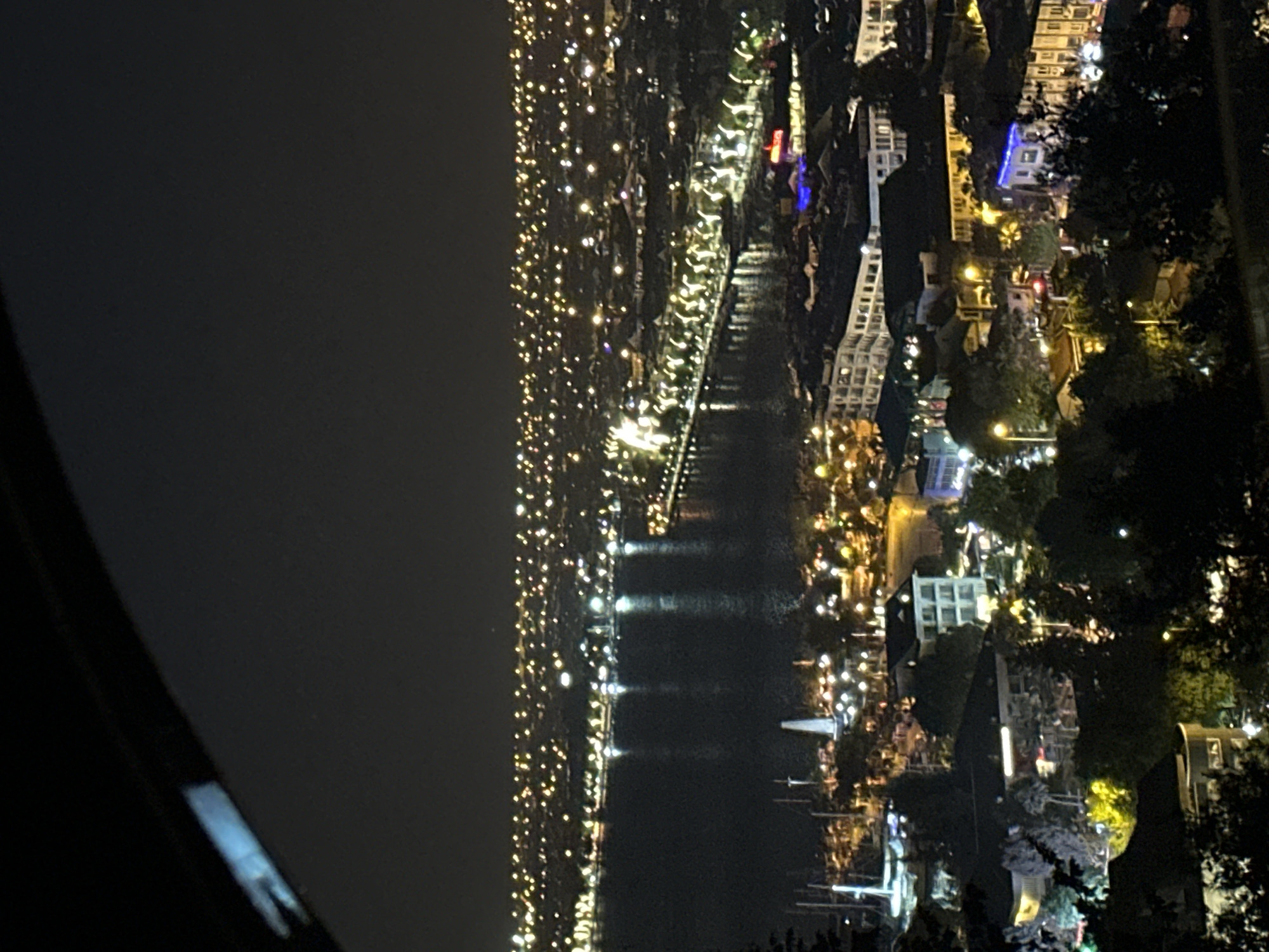 The image displays a vibrant night scene of a city built along a body of water, likely a bay or harbor, as seen from an elevated perspective. The time of day is clearly night, with a dark sky dominating the upper portion of the frame.
The scene is awash with countless artificial lights, primarily warm yellow and white, emanating from buildings and along the waterfront. These lights reflect distinctly on the calm surface of the water, creating long, shimmering vertical streaks.
On the closer landmass, which appears to be the right bank of the waterway, a dense array of structures, likely buildings and establishments, are illuminated. Interspersed among these are dark clusters of trees, indicating vegetation within the urban environment. A prominent, tall, and slender white structure, possibly a lighthouse or a monument, stands near the water's edge. Numerous boats, resembling yachts, are moored in the water nearby, adding their own lights to the reflections. Some blue and red lights are visible, hinting at specific businesses or decorative lighting.
Across the water, the opposing shore extends into the distance, marked by a sprawling tapestry of smaller, twinkling lights that signify a more expansive urban area. The overall impression is one of a lively, bustling city at night.
No specific activity or event is discernible, but the illuminated waterfront and presence of boats suggest a popular coastal or port city, consistent with the given location of Fethiye, Turkey. A dark, curved element in the bottom left foreground indicates the photograph was taken from behind a railing or window frame. No visible text is identifiable within the image.