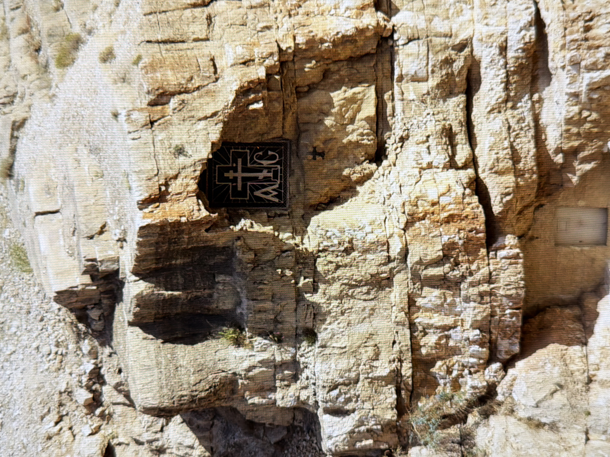 The image shows a section of a rocky cliff face. Carved into the rock is a dark rectangular opening, within which is a metal plaque displaying a Christian cross at the top and the letters "AIG" below. To the left of this opening, a small, dark cross is also carved into the rock. In the lower portion of the image, a rectangular doorway is visible, set into the base of the cliff. The surrounding environment is arid and appears to be a mountainous or hilly terrain with sparse vegetation. This scene is not located in Kyiv, Ukraine.