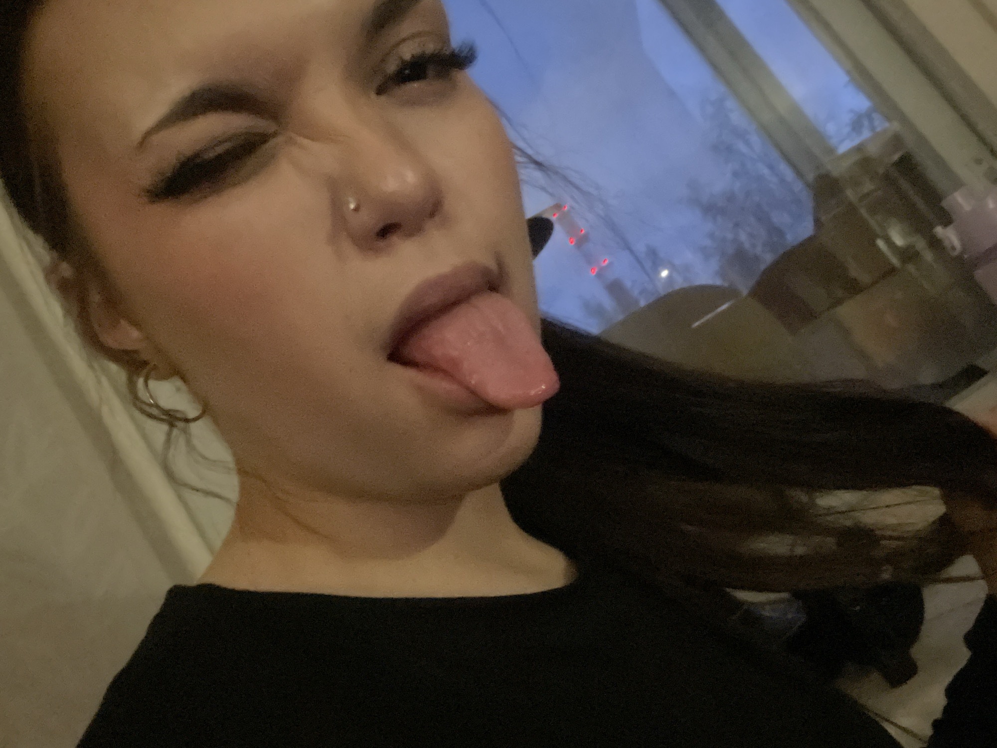A young woman is depicted in a close-up, eye-level shot, making a playful expression. She is winking with her left eye, sticking out her tongue, and slightly crinkling her nose. Her face is the central focus, featuring prominent dark eyebrows, long dark eyelashes (possibly false), and a small silver stud piercing on her left nostril. Her dark hair is visible, swept to her right side, and a silver hoop earring can be seen on her left ear. She is wearing a simple black top.
The background is out of focus, revealing a window looking out onto an outdoor scene. The sky appears a deep blue, indicative of dusk or twilight, with bare tree branches silhouetted against it, suggesting a colder season. Distant red lights, possibly from vehicles or city infrastructure, are visible through the window, implying an urban or suburban setting. The overall lighting is dim, with some ambient light entering from the window. The scene is consistent with an indoor setting in an urban environment like Saint Petersburg, Russia, as provided in the context. There is no visible text.