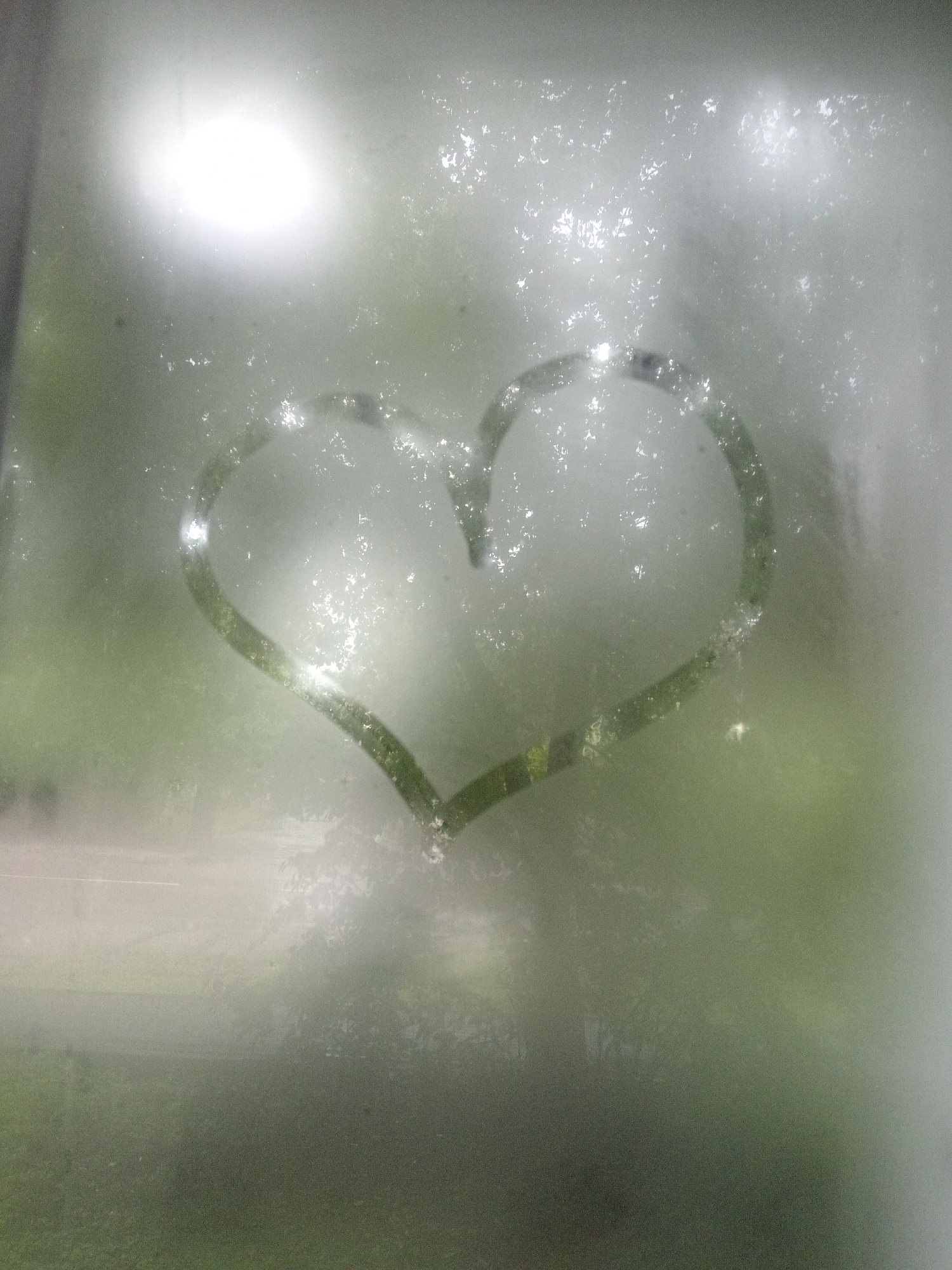 A heart is drawn on a steamy window. The heart shape is defined by clear lines against the condensation. Outside the window, a blurry green landscape is visible, suggesting an outdoor environment. The light source is bright and diffused, creating a halo effect and reflecting off the water droplets on the glass. The image appears to be taken in Belogorsk, Russia, as per the prompt's request, although there are no specific visual cues within the image to confirm this location. The scene is static and lacks any human presence or action beyond the drawing of the heart.
