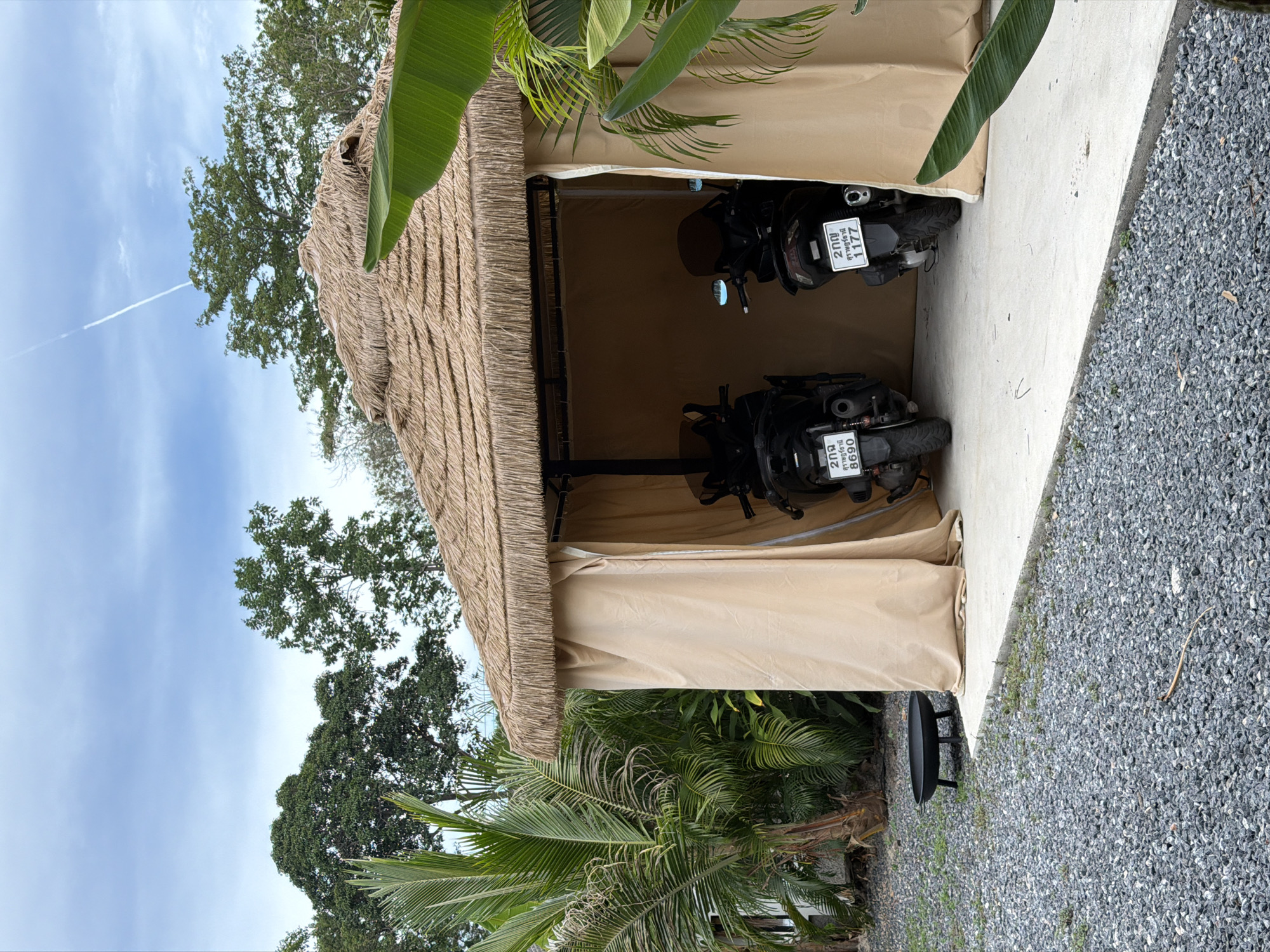 In Baan Bo Phut, Thailand, a thatched-roof shelter with beige fabric walls stands on a concrete pad. Two black scooters are parked inside the shelter, facing forward. The scooter on the left has a license plate that reads "29890" and the scooter on the right has a license plate that reads "21177". A black metal fire pit with three legs is situated on a gravel-covered ground to the left of the shelter. Tropical plants, including palm trees, surround the scene. The sky above is partly cloudy with a faint white contrail.