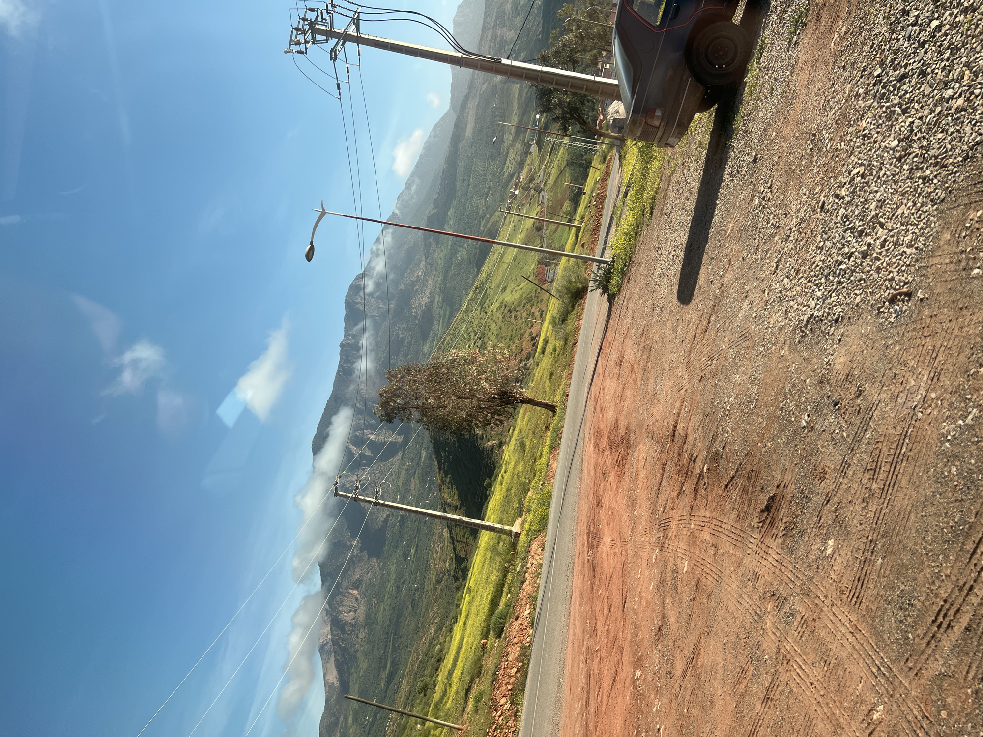The image shows a scene near Ouaouizert, Morocco. The foreground features a dirt and gravel road with tire tracks. A portion of a dark-colored vehicle is visible on the right side of the frame. Beyond the road is a green hillside with scattered vegetation and yellow flowers. In the mid-ground, utility poles and power lines run along the hillside. A single tree stands in the middle of the hillside. In the background, mountains are partially obscured by clouds. The sky is blue with some cloud cover.