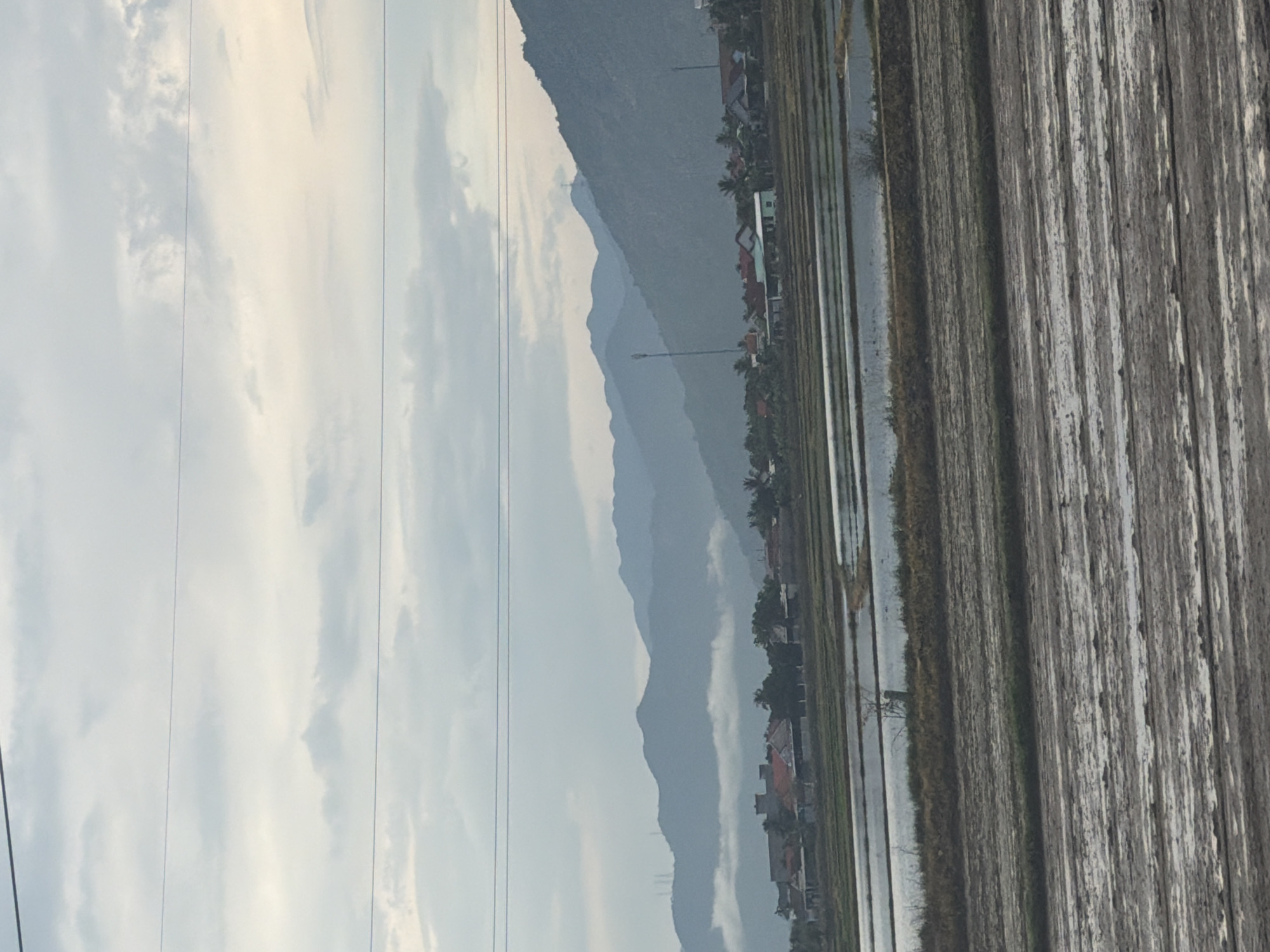 A wide-angle, outdoor, eye-level shot captures a rural landscape in Dien Lac, Vietnam, under an overcast sky. In the foreground, newly plowed fields lie dormant, appearing muddy and wet with visible furrows. Beyond these fields, several rectangular rice paddies are flooded, reflecting the muted light of the sky. A thin line of dry vegetation separates the plowed fields from the paddies. In the mid-ground, a collection of houses with red-tiled roofs forms a small village, nestled among sparse trees. Two prominent cellular towers stand out amidst the houses. In the background, a series of undulating mountains stretches across the horizon, partially obscured by a layer of low-lying clouds that hug the foothills. The overall atmosphere is serene and quiet, suggesting an early morning or late afternoon scene.