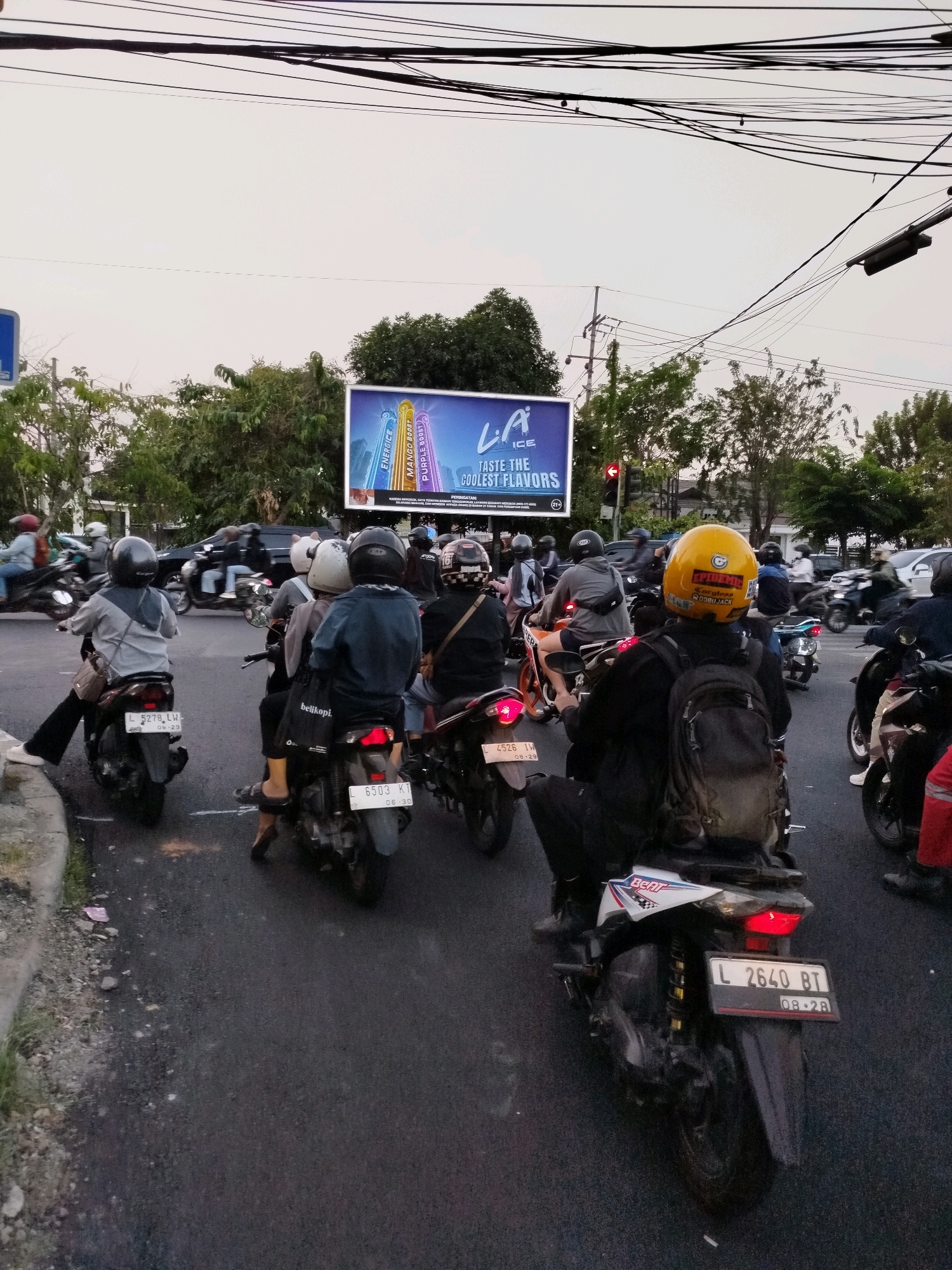 The image depicts a traffic scene in Surabaya, Indonesia, with several motorcycles and their riders on a paved road. The sky is overcast, suggesting it may be late afternoon or early morning. A large billboard advertising "L.A. ICE" with the tagline "TASTE THE COOLEST FLAVORS" is visible in the background, along with trees and utility poles. The motorcycles are in motion or stopped in traffic, with riders wearing helmets for safety. Some of the motorcycle license plates are visible, showing alphanumeric combinations common in Indonesia. The overall impression is a snapshot of everyday urban life and transportation in a bustling Indonesian city.