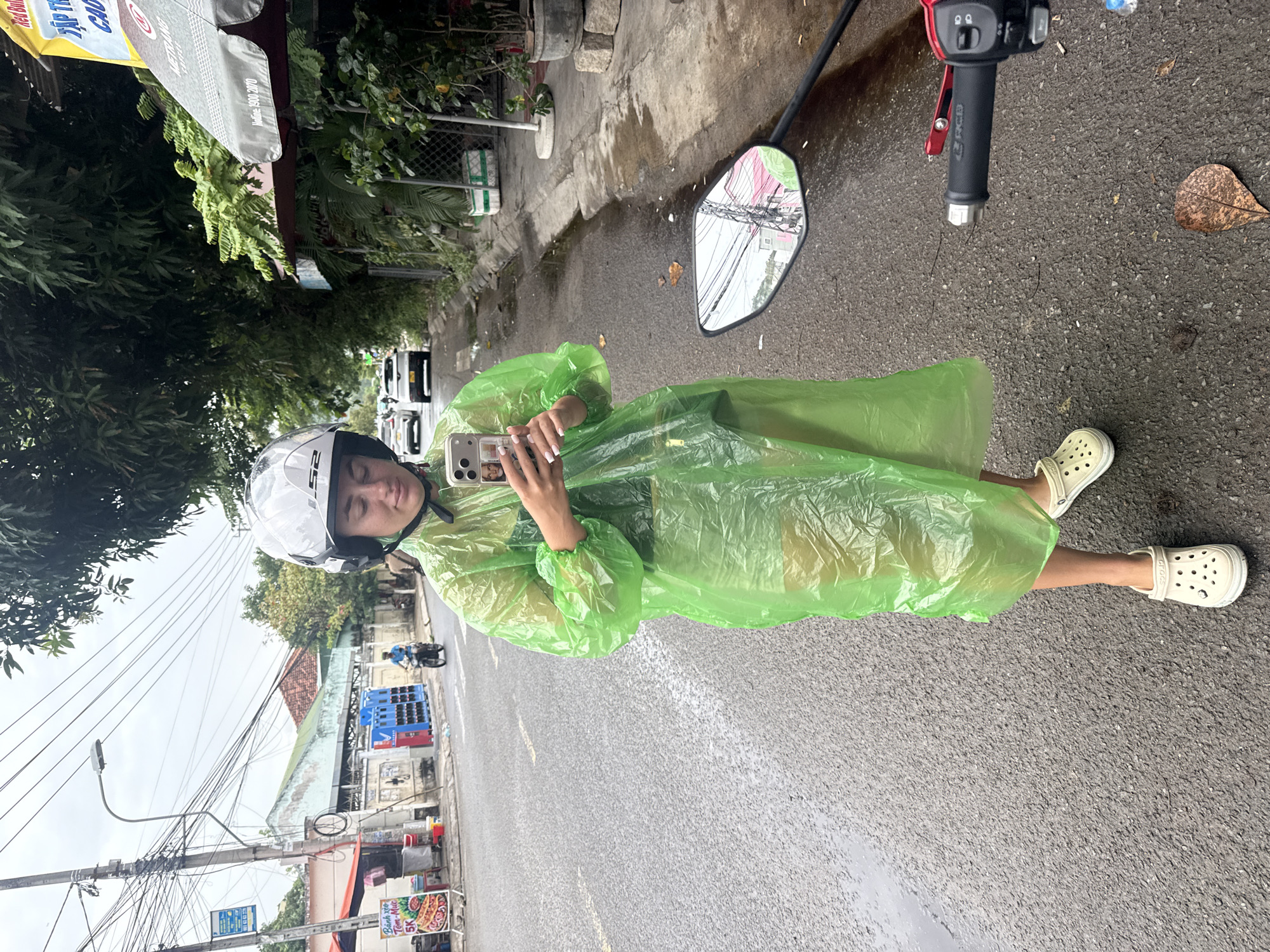 In Nha Trang, Vietnam, a person stands on a wet asphalt road wearing a bright green, translucent raincoat, a white helmet with a black strap, and white Crocs. The person is holding a smartphone and appears to be taking a selfie. To the right, the handlebar and side mirror of a red motorcycle are visible. In the background, there are trees, power lines, buildings, and parked cars. The sky is overcast. A street vendor's stall with a menu board advertising food is visible on the left side of the road.