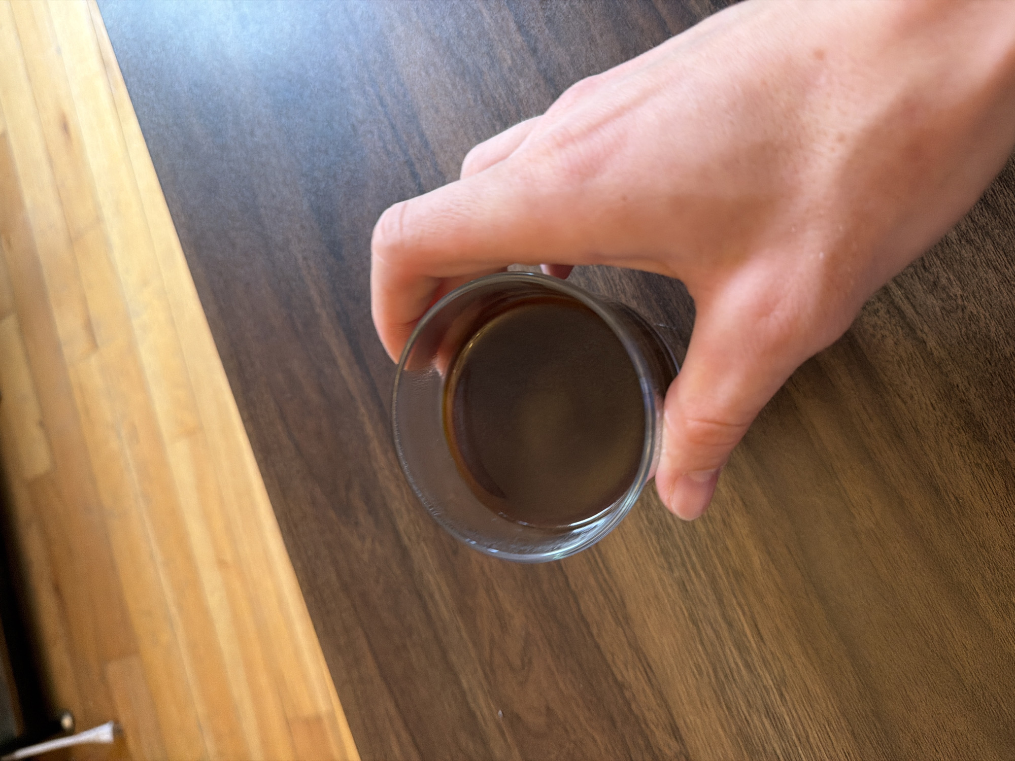 A hand grips a glass of dark liquid on a brown wooden surface. The hand is pale and the glass is clear. To the left of the surface is a lighter wooden surface. No indication of location.