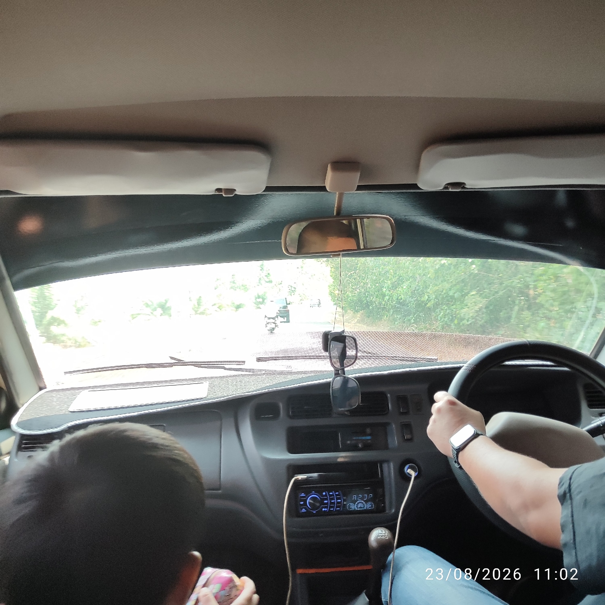 The image shows the interior of a car, viewed from the perspective of a passenger. A person's arm and hand are visible on the right, resting on the steering wheel. The person is wearing a watch. In the foreground, on the left, the head and shoulder of a child are visible. The child is holding a pink object. The dashboard of the car is visible, with a car stereo and a gear shift. Outside the car, through the windshield, a road is visible, with trees on either side. There is another vehicle in the distance on the road. The timestamp at the bottom right corner of the image indicates "23/08/2026 11:02". This scene is not specifically identifiable as being in Kacang Botor, Indonesia.