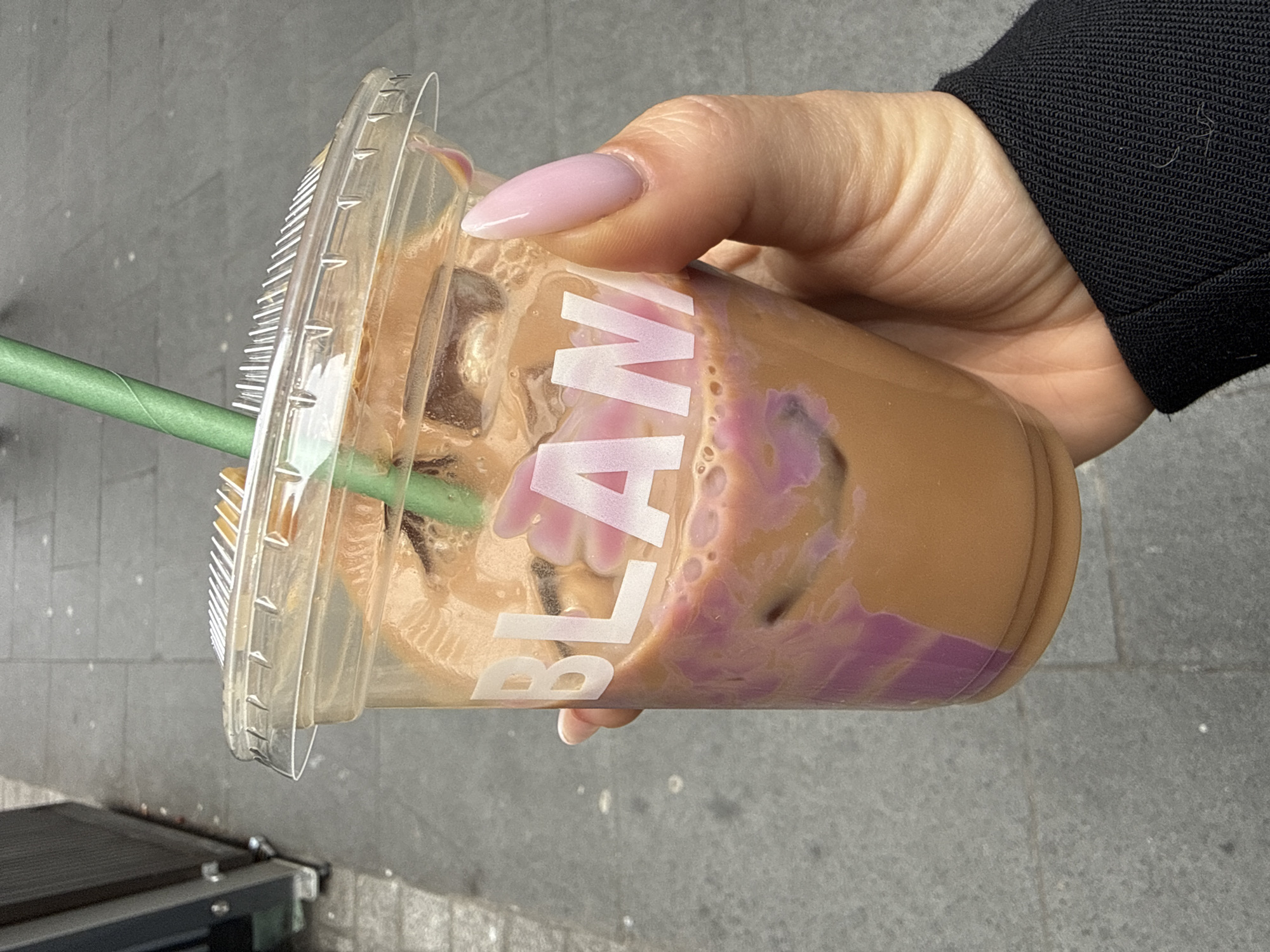 The image presents a close-up, slightly high-angle view of a person's hand holding a clear plastic beverage cup over a paved pedestrian surface in the City of London, United Kingdom. The cup contains a layered iced beverage, consisting primarily of a light brown liquid and distinctive swirls of light purple liquid, with several ice cubes visible. A green paper-like straw is inserted through the clear plastic domed lid. White sans-serif lettering, partially visible as 'BLAN' followed by a partial letter, is printed on the cup's exterior. The visible nails on the hand are manicured, featuring a light pink polish and an almond shape. The wrist is partially covered by a black long-sleeved garment. The background consists of an out-of-focus grey paved ground, consistent with a sidewalk, and a dark metallic structure with visible hinges or bolts positioned to the far left.