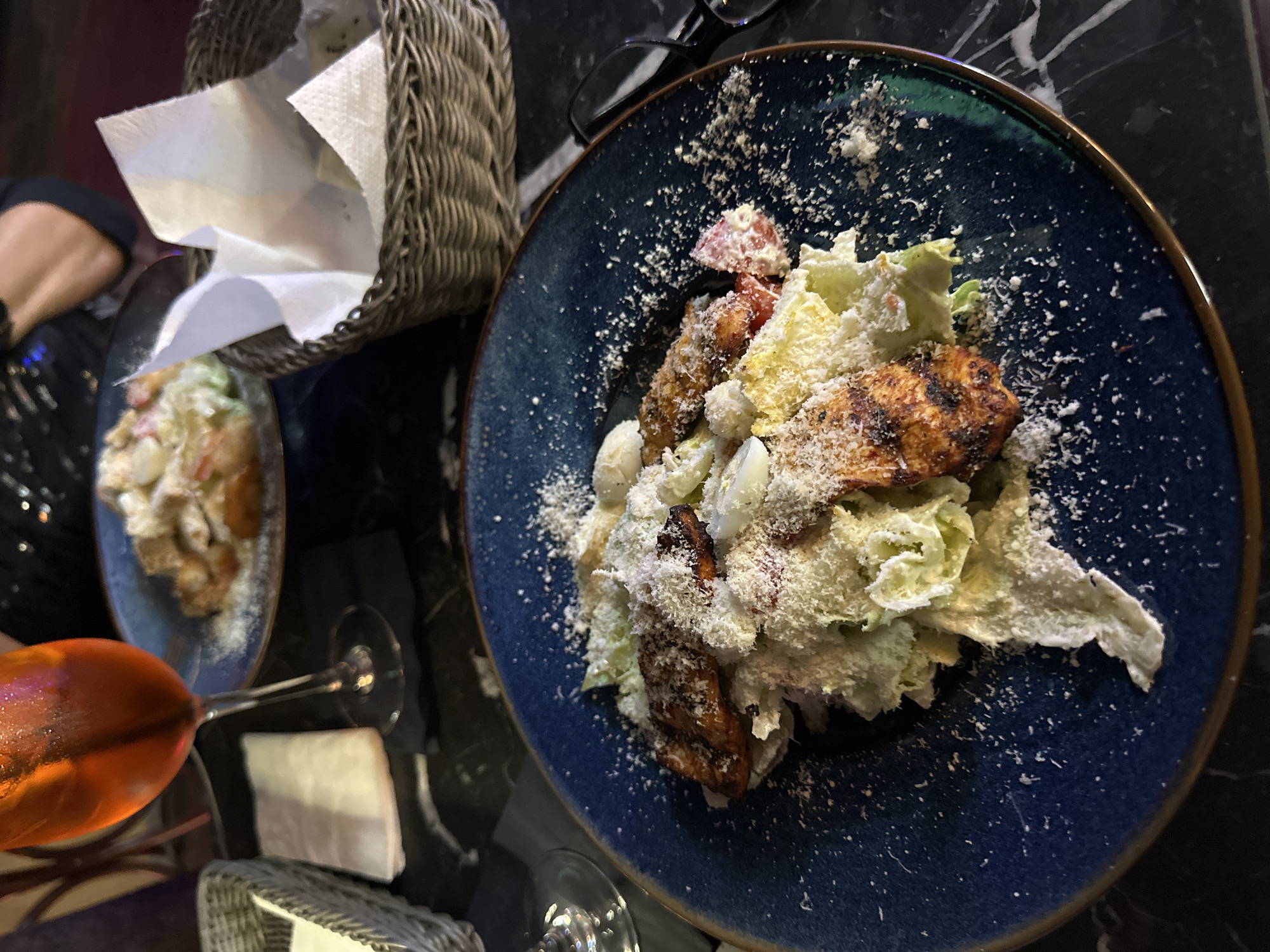 The image shows a restaurant table in Wrocław, Poland, featuring two plates of Caesar salad, a basket of napkins, and a drink. The Caesar salad on the right is presented on a blue plate, garnished with lettuce, chicken, croutons, eggs, tomatoes, and parmesan cheese. There is another Caesar salad plate on the left, along with a bright orange-colored drink and glasses. A basket filled with white napkins is placed near the upper-left corner. Black marble table. The photo appears to be taken from an overhead perspective.