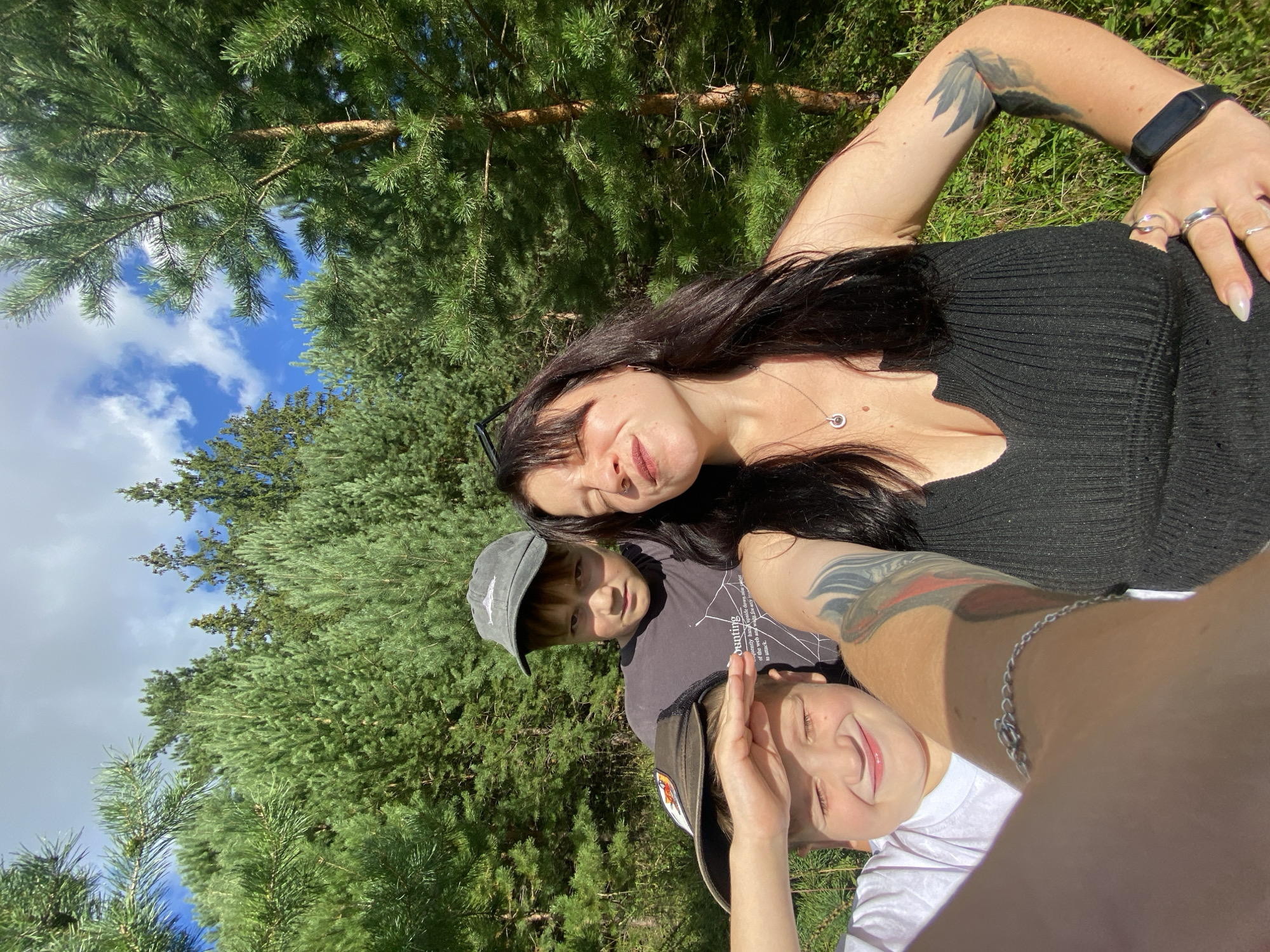 In the city of Fokino, Russia, on a sunny day, a woman and two boys are captured in a selfie taken outdoors. The background is dominated by a dense forest of coniferous trees. The woman, with long dark hair and bangs, is positioned in the foreground on the right. She is wearing a black ribbed top with a deep V-neck and has her right hand on her hip. She is adorned with several rings and a smartwatch on her left wrist, and also sports a bracelet on her left arm, which extends into the frame. Tattoos are visible on her arms. The two boys are positioned to her left and in the center, both wearing baseball caps. The boy on the far left is wearing a white t-shirt and has his eyes closed and a smile on his face, with his hand shading his eyes. The boy in the center is wearing a dark t-shirt and a gray baseball cap, looking directly at the camera with a neutral expression. The overall impression is a casual outdoor family moment.