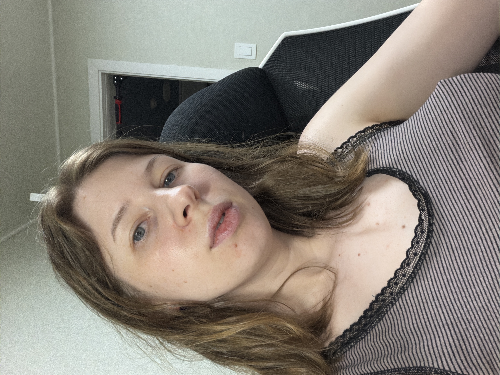 A young woman with shoulder-length brown hair, fair skin, and blue eyes is captured in a selfie-style shot, seated in a black and white office chair. She is wearing a horizontally striped tank top with black lace trim. The background appears to be an indoor setting with a plain wall and a doorway in the upper right corner, possibly in Dongducheon-si, South Korea.