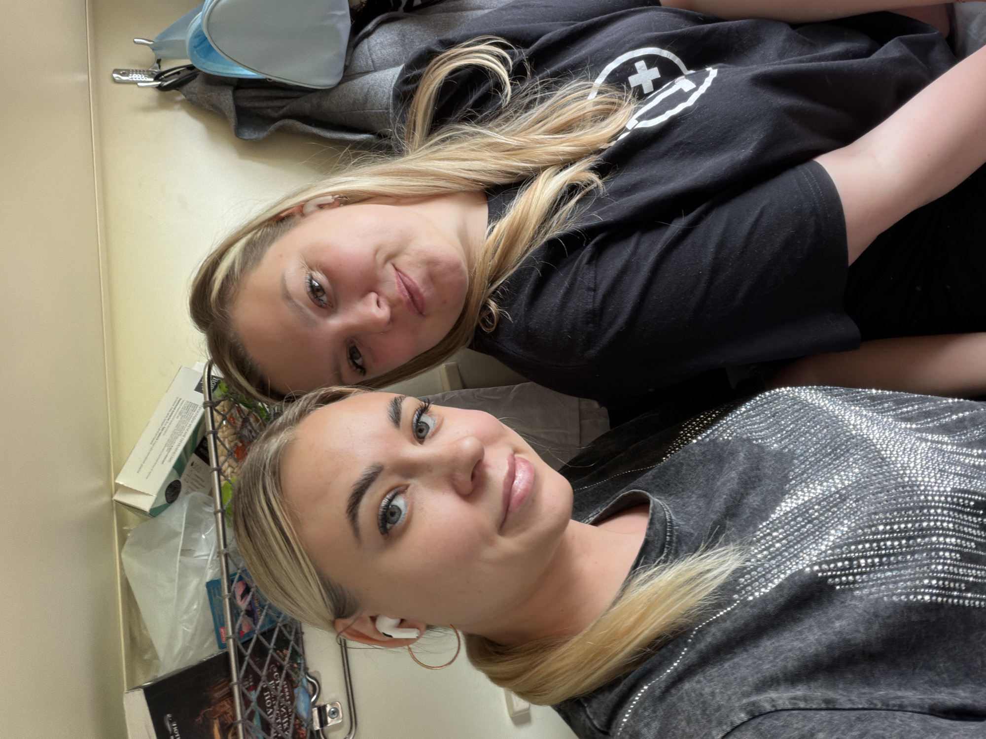 Two young Caucasian women are posing for a selfie in an indoor setting. The woman in the foreground is wearing a dark gray shirt with rhinestone embellishments, and she has blonde hair pulled back into a low ponytail with a gold hoop earring and a white earbud in her left ear. The woman in the background is wearing a black T-shirt with a white circular logo that contains a plus sign. She has blonde hair with darker roots. A metal wire basket can be seen to the left of the frame, containing various items including a box, a plastic bag, and books. A gray garment and a light blue bag are in the background. The photo appears to have been taken inside, perhaps on a bus or train, given the limited space and arrangement of objects. There is no verifiable information to suggest the location is in the city of Aksay, Russia.