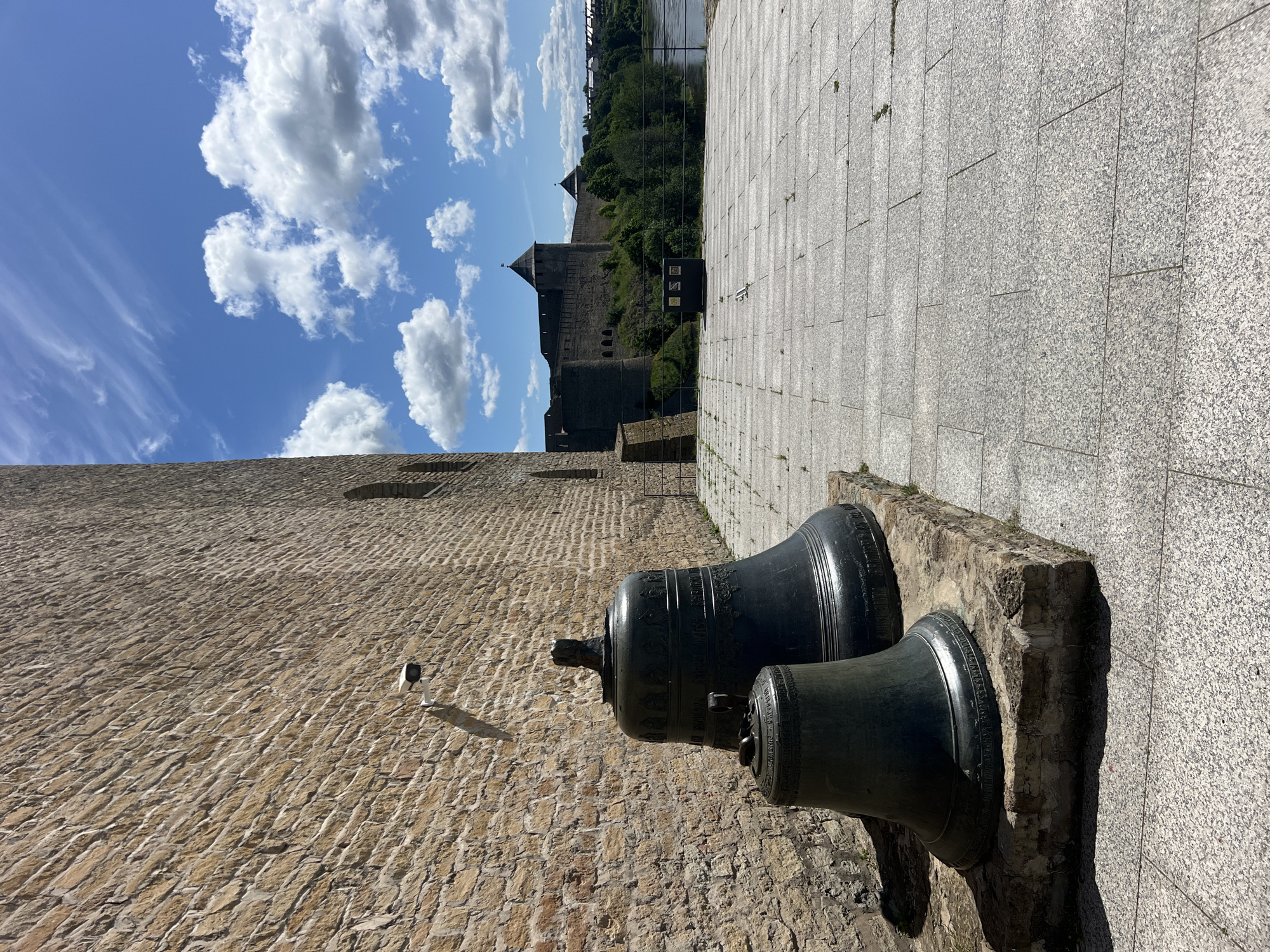 This image captures a view of a stone wall and two large bells in the foreground, with a medieval-style castle and a river in the background. The sky is a vibrant blue with fluffy white clouds, suggesting a clear and sunny day. The stone wall in the foreground is constructed from rough-hewn, tan-colored stones, laid in a rustic pattern. Two large, dark-colored bells are prominently displayed on a stone pedestal. They appear to be old and possibly decorative, with intricate designs visible around their rims.
Beyond the bells, a paved walkway made of large, light-grey stone tiles stretches into the distance. To the left of the walkway, a massive stone structure, possibly part of a fortification or historical building, rises up. Further in the background, a substantial stone castle with towers and battlements sits atop a hill, surrounded by lush green trees. A body of water, likely a river, is visible to the right of the castle, with some structures on the opposite bank.
The overall scene evokes a sense of history and grandeur. The bright daylight and blue sky contribute to a pleasant and inviting atmosphere. The presence of the castle and the ancient bells suggests a historical site or museum. While no people are visible in the image, the well-maintained walkway and the presence of what appears to be a security camera on the stone wall indicate that this is an accessible public space. The orientation of the image is such that it appears to be taken from a low angle, looking upwards towards the castle and sky.