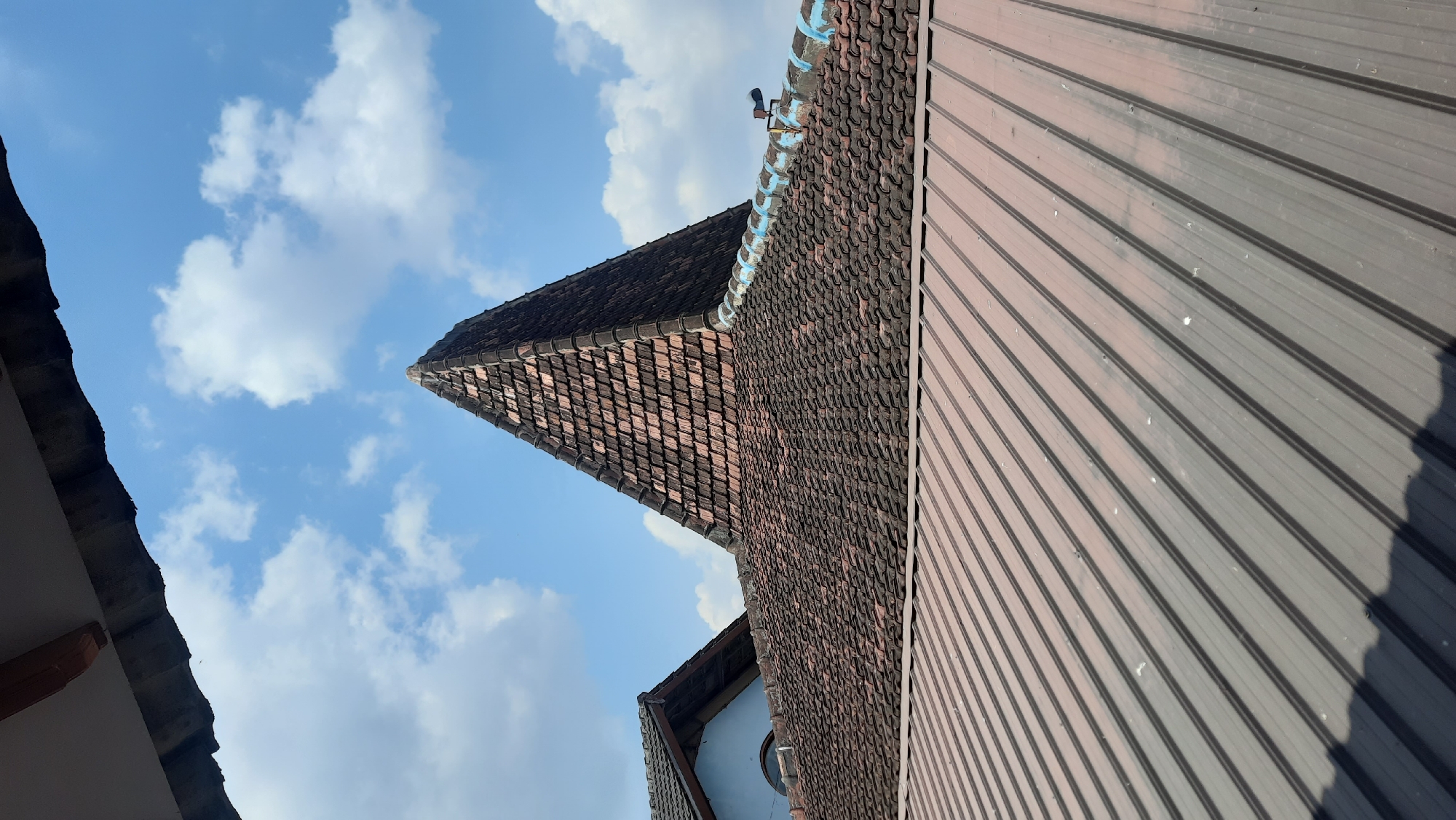 The image shows a view of rooftops under a partly cloudy sky. In the foreground, a corrugated metal roof with parallel ridges is visible, casting a shadow on the left. Behind this, an older roof covered in dark, overlapping tiles forms a curved surface. Further back, a prominent pyramid-shaped roof constructed with reddish-brown tiles dominates the center of the frame. To the left of the pyramid, another tiled roof is partially visible, with a blue wall and a curved architectural element. The sky above is a clear blue with scattered white cumulus clouds. The scene appears to be an outdoor environment, possibly residential, given the roofing materials. There are no people or animals visible in the image.
