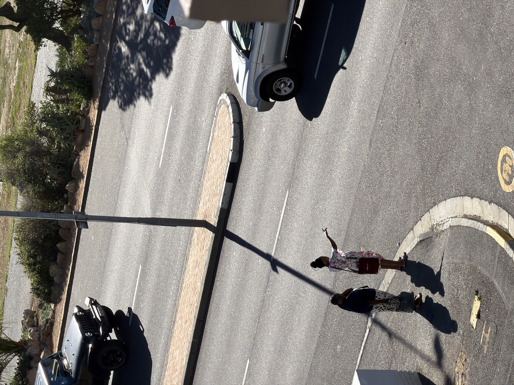 An aerial view of a street in Cape Town, South Africa, shows two people standing on the road near a curb. One person is wearing a floral dress and holding their arm up in the air. The other person is wearing a dark shirt and patterned pants. A silver car is parked on the opposite side of the street, and a dark-colored Jeep is parked at the lower left. A lamppost casts a shadow across the street. The road surface is asphalt with white lane markings. A brick-paved median runs along the street's center.