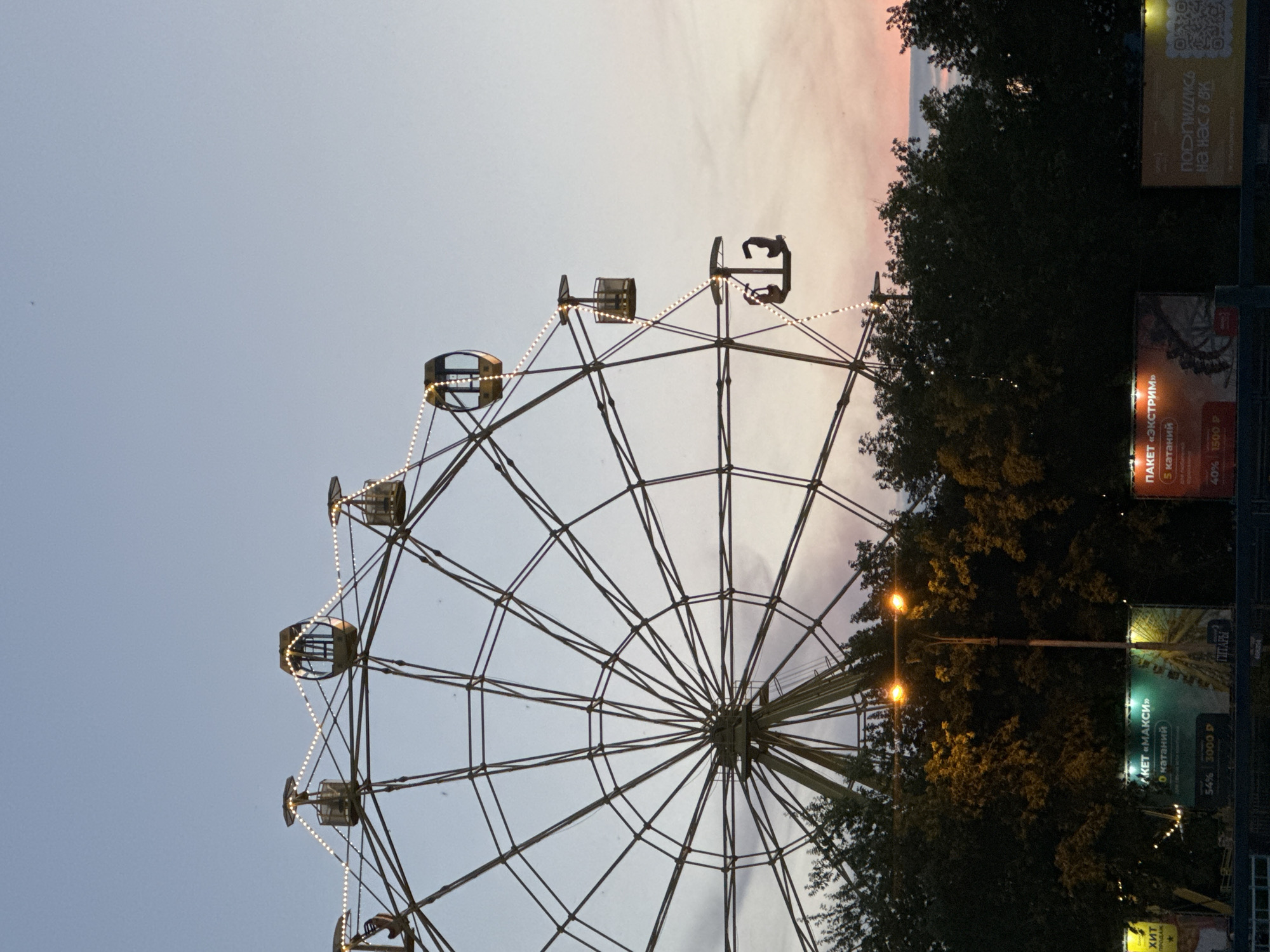An exterior, vertical, eye-level shot captures a Ferris wheel in the city of Unknown, Unknown, during twilight. The Ferris wheel dominates the frame, its metal structure silhouetted against a pale, gradient sky that transitions from a soft blue at the top to a warm pink and orange hue near the horizon. String lights are draped along the spokes of the Ferris wheel, their individual bulbs creating a festive sparkle. Several gondolas are visible, some empty and others occupied by a silhouette of a person or two, suggesting they are in motion. The Ferris wheel is set against a backdrop of dark, lush trees, some of which are illuminated by streetlights below. In the foreground, glimpses of billboards with Russian text are visible, further indicating an urban setting. The overall atmosphere is serene, with the soft lighting of twilight and the gentle glow of the Ferris wheel creating a peaceful scene.