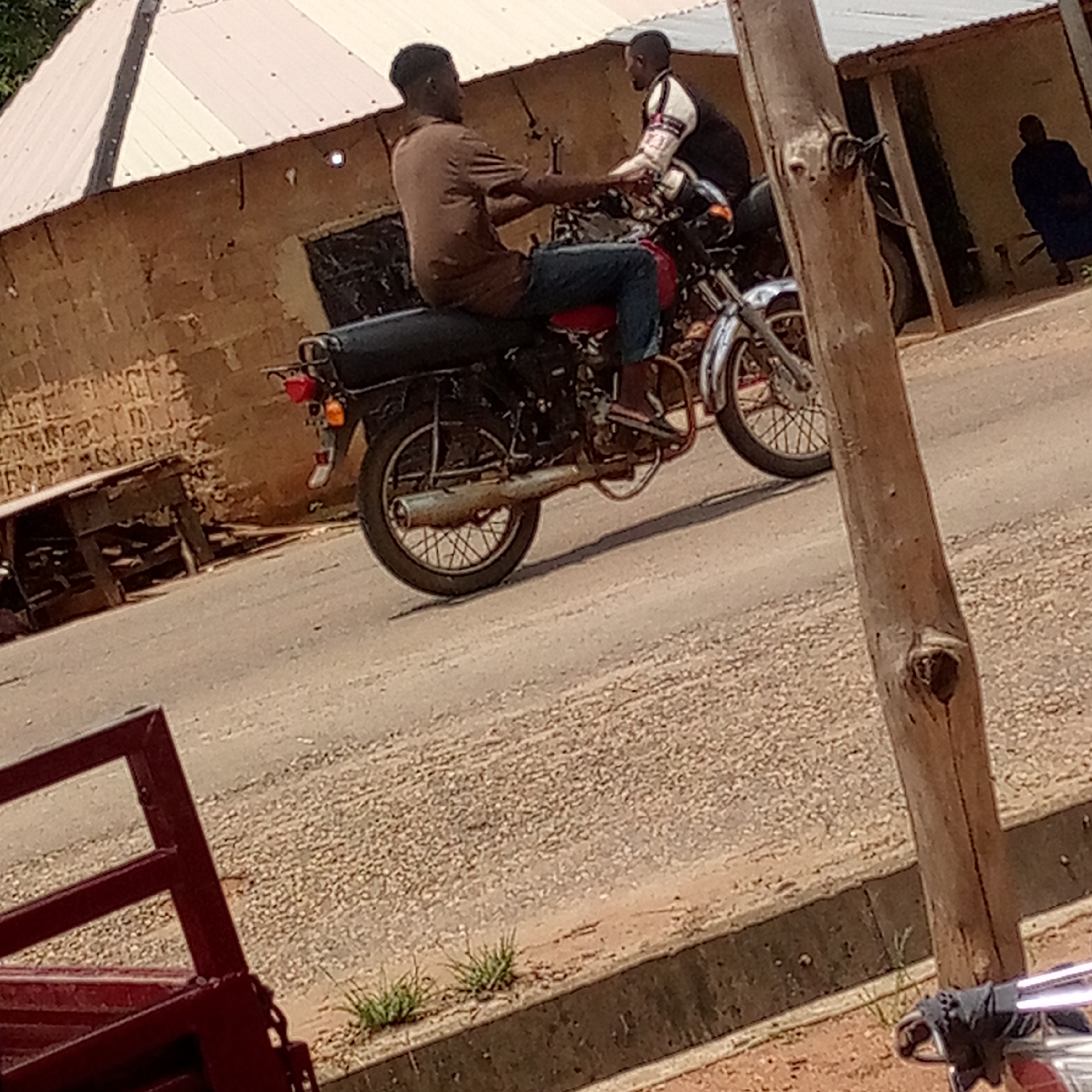 The image depicts an outdoor scene in what appears to be a semi-urban or rural area, consistent with Mararaba, Nigeria. Two men are prominently featured on motorcycles, seemingly interacting on a dusty, gravelly road.
The man in the foreground is seated on a dark motorcycle with a red seat, wearing a brown short-sleeved shirt and blue jeans. He is positioned facing left but looking towards the right. Slightly behind him and to the right, a second man is also on a motorcycle, wearing a white and black jacket, and appears to be facing or looking towards the first man. Both motorcycles are standard models, with visible chrome elements and exhaust systems.
In the background, a single-story building with walls made of light brown, unpainted bricks or mud bricks and a corrugated metal roof stretches across the scene. To the far left, a wooden table or stand is placed against the building wall. To the right side of the building, a third man, dressed in a dark blue or black garment, is visible near a doorway.
The ground is unpaved, consisting of dirt and small gravel, with scattered small clumps of green grass growing along a concrete curb or drainage channel. A thick, reddish-brown wooden pole stands prominently in the foreground on the right, partially obscuring the second motorcycle. In the bottom left corner, a dark red metal frame object is partially visible. The scene is illuminated by natural daylight, suggesting it is daytime. There is no visible text in the image.