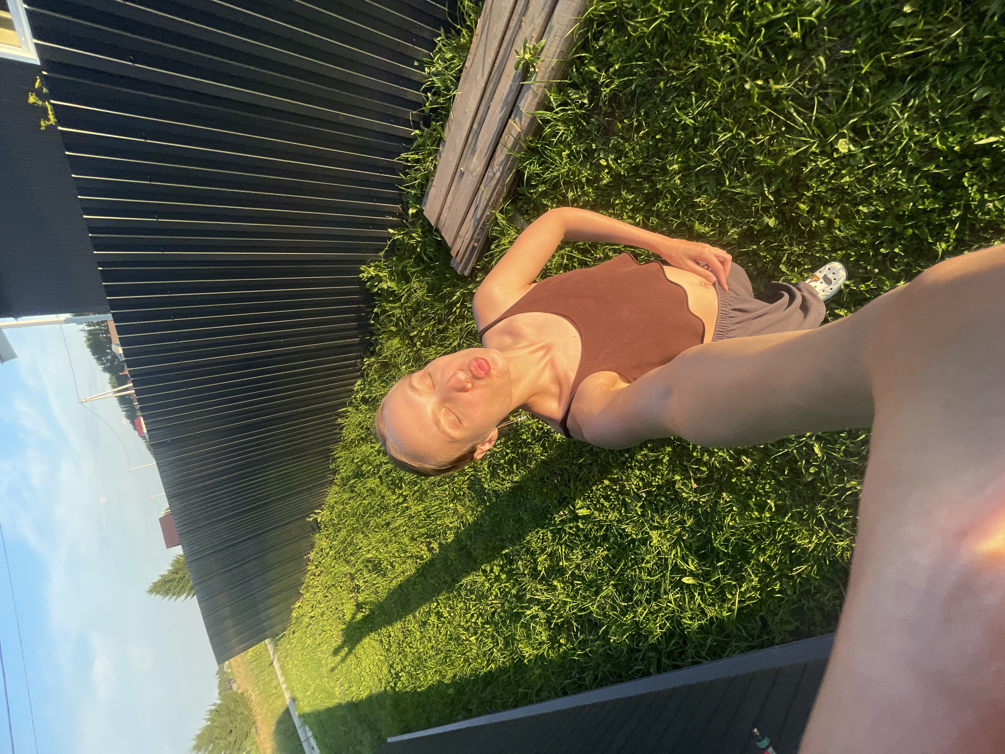 The image was taken in the city of Podshivalovo, Russia. A young woman is standing on a grassy lawn in front of a dark metal fence. She is wearing a brown tank top and gray sweatpants, and white crocs. She is holding her phone out to take a selfie, with her eyes closed and her lips puckered as if blowing a kiss. The sun is setting, casting long shadows across the grass. In the background, there are trees and houses. The scene appears to be a residential area.