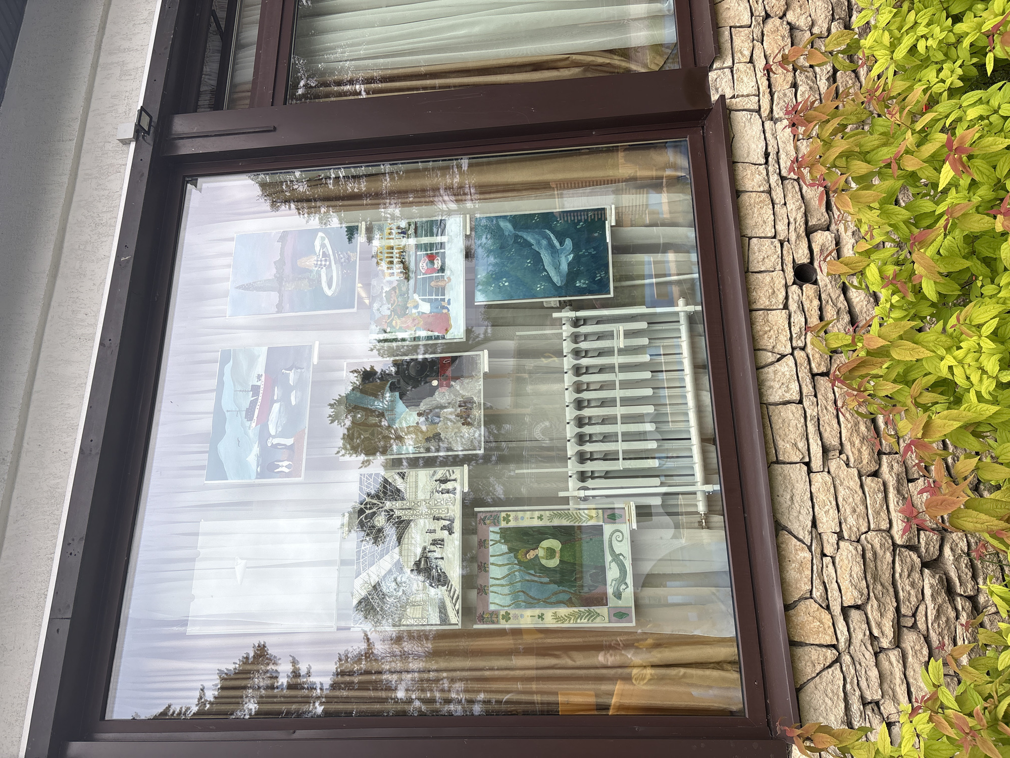 The image shows a display of artwork in a large window of a building. The artwork includes several framed prints, some depicting ships and icebergs, others featuring people in various settings, and one with a shark. There is also a radiator visible behind some of the prints. The window is framed by dark brown trim and overlooks a stone wall with some greenery in the foreground. The overall scene suggests a public display of art, possibly in a cultural center or gallery in Sestroretsk, Russia.
