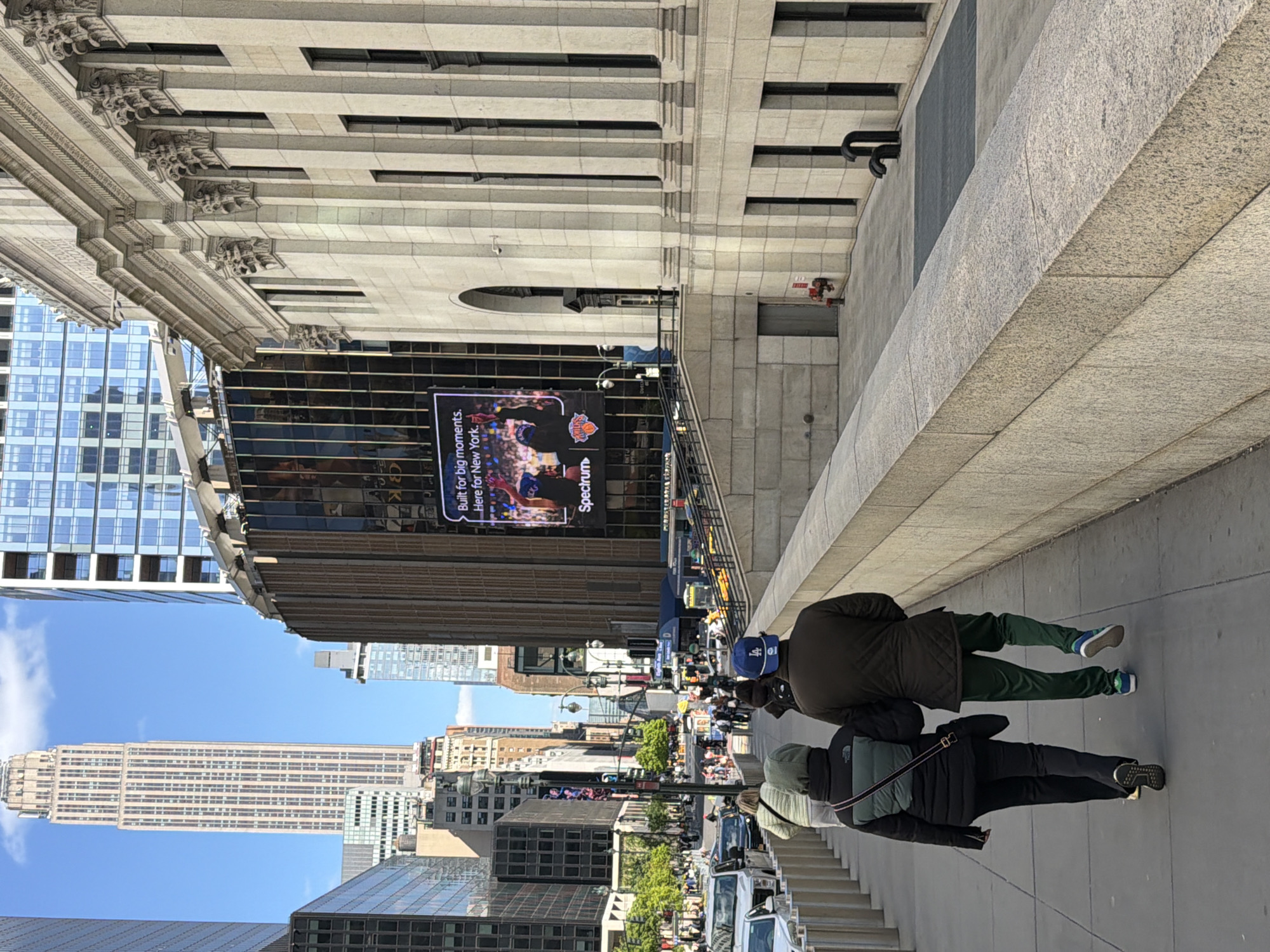 This photograph depicts a street scene in New York City, United States of America. In the distance, the Empire State Building is visible. Madison Square Garden can be seen with a digital sign advertising the New York Knicks basketball team. There are people walking on a sidewalk in the foreground. The architectural details of a building are visible on the left and top of the image. Cars are parked along the street.