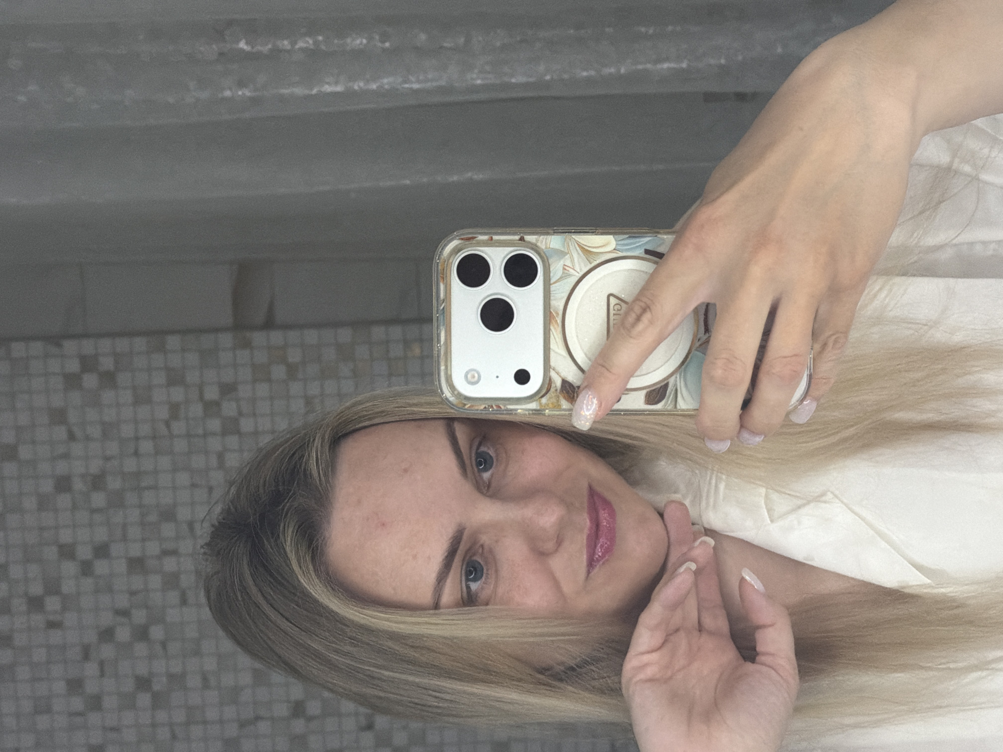 This mirror selfie, captured in Votkinsk, Russia, features a blonde woman with blue eyes and fair skin. She is positioned in the foreground, with her head slightly tilted and a subtle, pleasant expression on her face, accented by pink lipstick. Her blonde hair cascades around her shoulders.
The woman holds a smartphone in her right hand, which has a clear case adorned with a light-colored floral or abstract pattern. A round pop socket or phone grip is attached to the back of the phone case, displaying the partial word "GIRL" in a serif font. Her fingernails on both hands are manicured with a light, possibly glittery or pearlescent polish. Her left hand rests gently near her chin.
The background, visible as a reflection in the mirror, consists of a wall covered in small, light-toned square tiles, suggesting an indoor setting like a bathroom or a tiled hallway. Above the tiled section, a blurred area indicates a shelf or another wall. The photo captures a moment of self-portraiture.