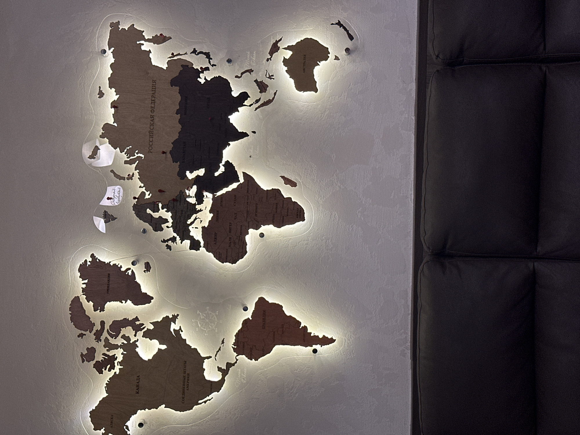 The image shows a wooden world map mounted on a textured wall above a dark leather sofa. The map is illuminated from behind with warm LED lights, creating a halo effect around the continents. Several small red pins are visible on the map, marking various locations. In the top center of the map, two small white paper flags are attached, one with handwritten text in Russian. The overall impression is of a decorative and functional piece of wall art. There is no indication of the city of Yuzhnyye Koryaki, Russia in the image, nor any people or actions taking place.