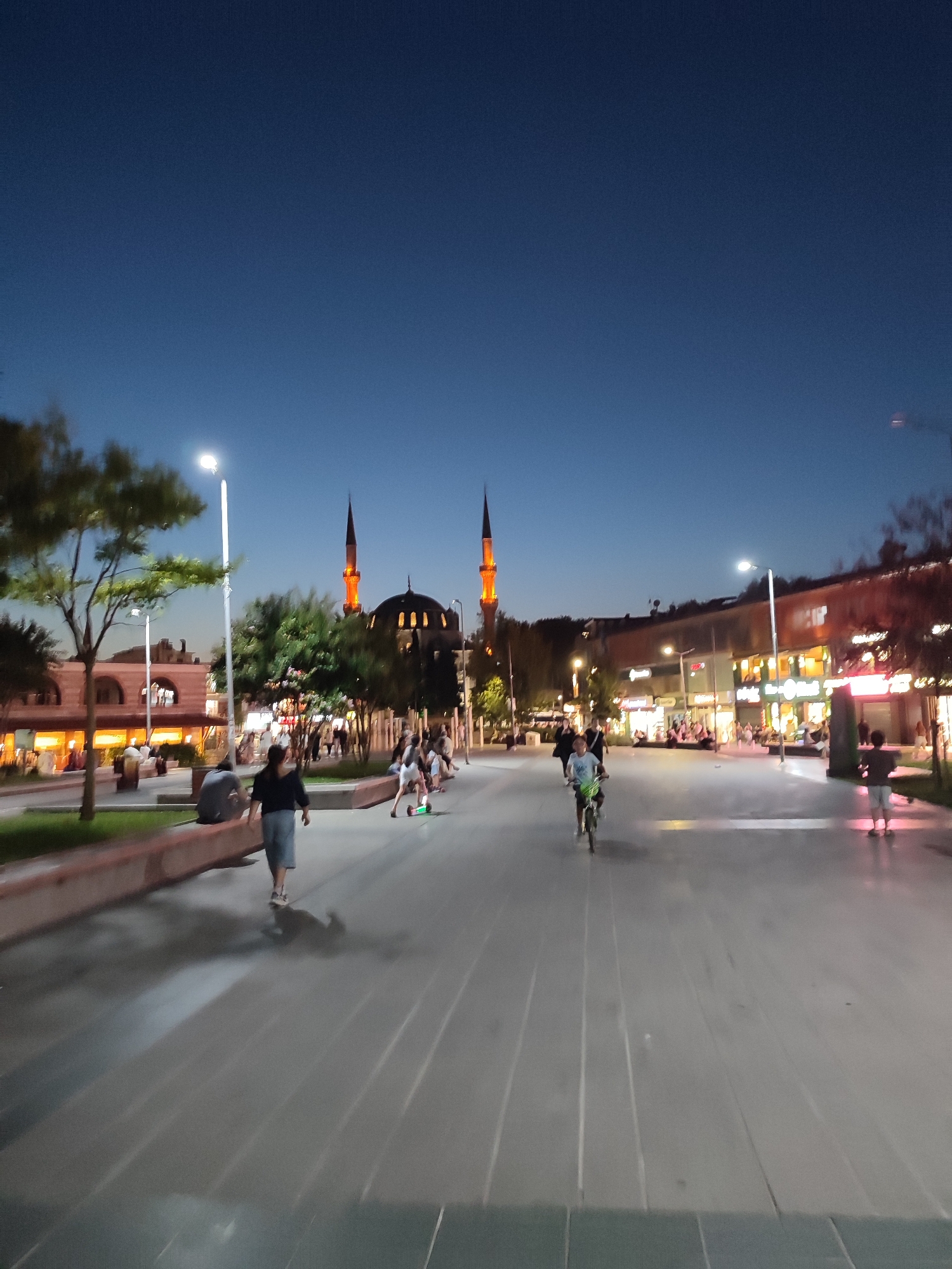 The image depicts a street scene in Üsküdar, Turkey, at dusk. A large mosque with two illuminated minarets is prominently visible in the background. The sky is a deep blue, indicating the transition from daylight to night. Several people are present in the scene, with some walking and others engaged in recreational activities. One individual is riding a bicycle, and another is on a scooter. The street is paved, and there are trees and streetlights lining the walkway. Buildings with illuminated windows line the street on the right side, suggesting the presence of shops or restaurants. The overall atmosphere appears to be one of a lively urban environment in the evening.